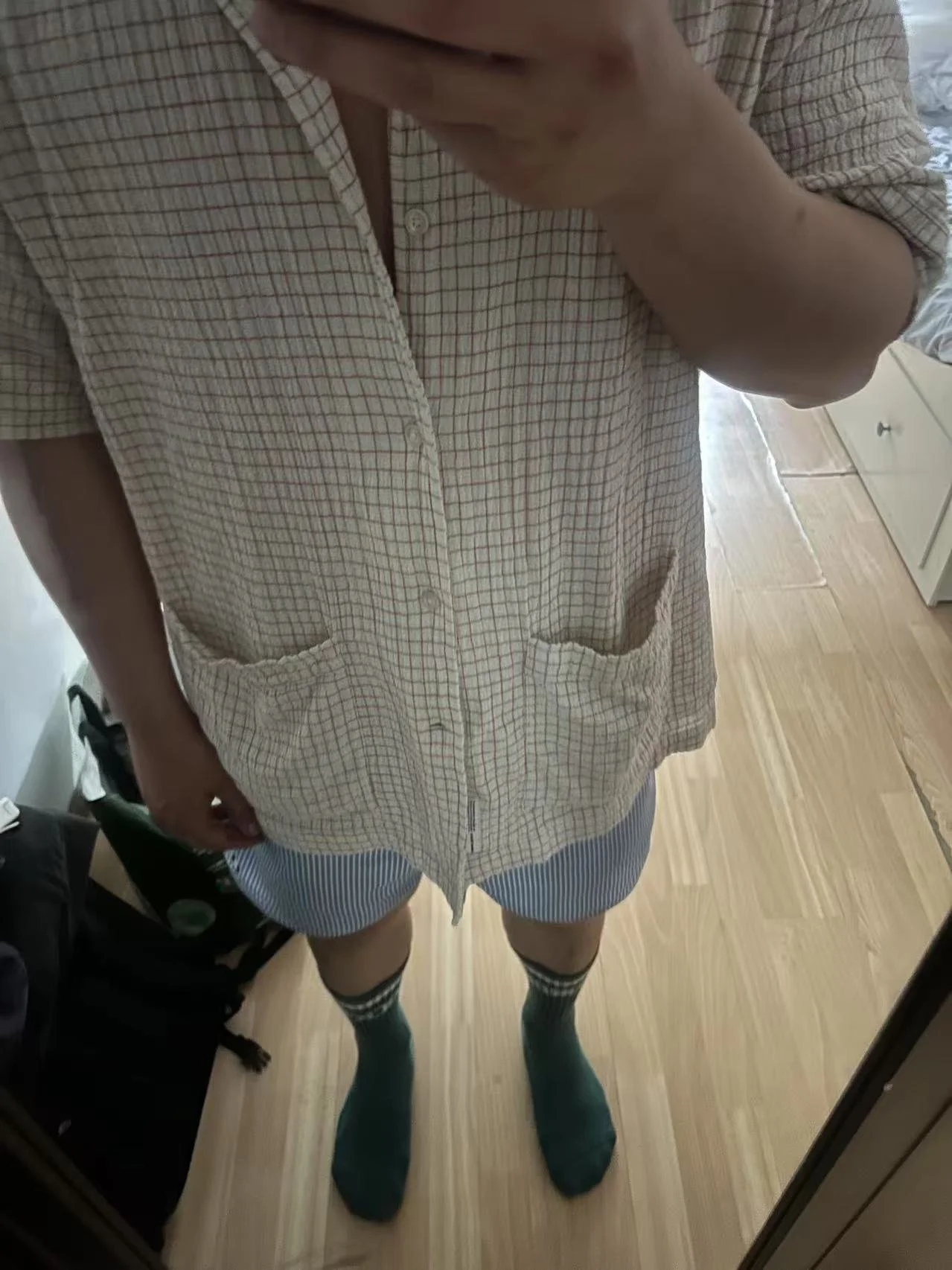 Person taking a mirror selfie. The person is wearing a checkered short-sleeve shirt, striped shorts, and green knee-high socks.