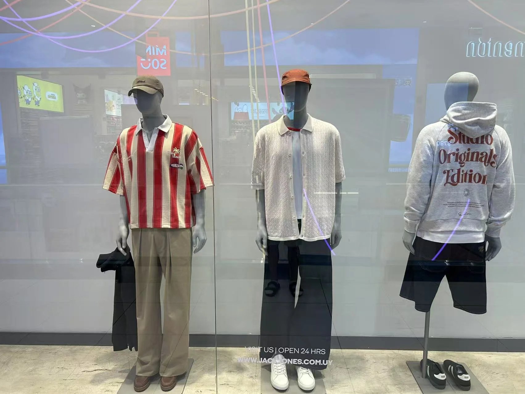 Three mannequins dressed in casual clothing are displayed in a storefront window. The mannequin on the left wears a striped short-sleeve shirt and khaki pants with a baseball cap. The middle mannequin wears a white textured short-sleeve shirt, dark p