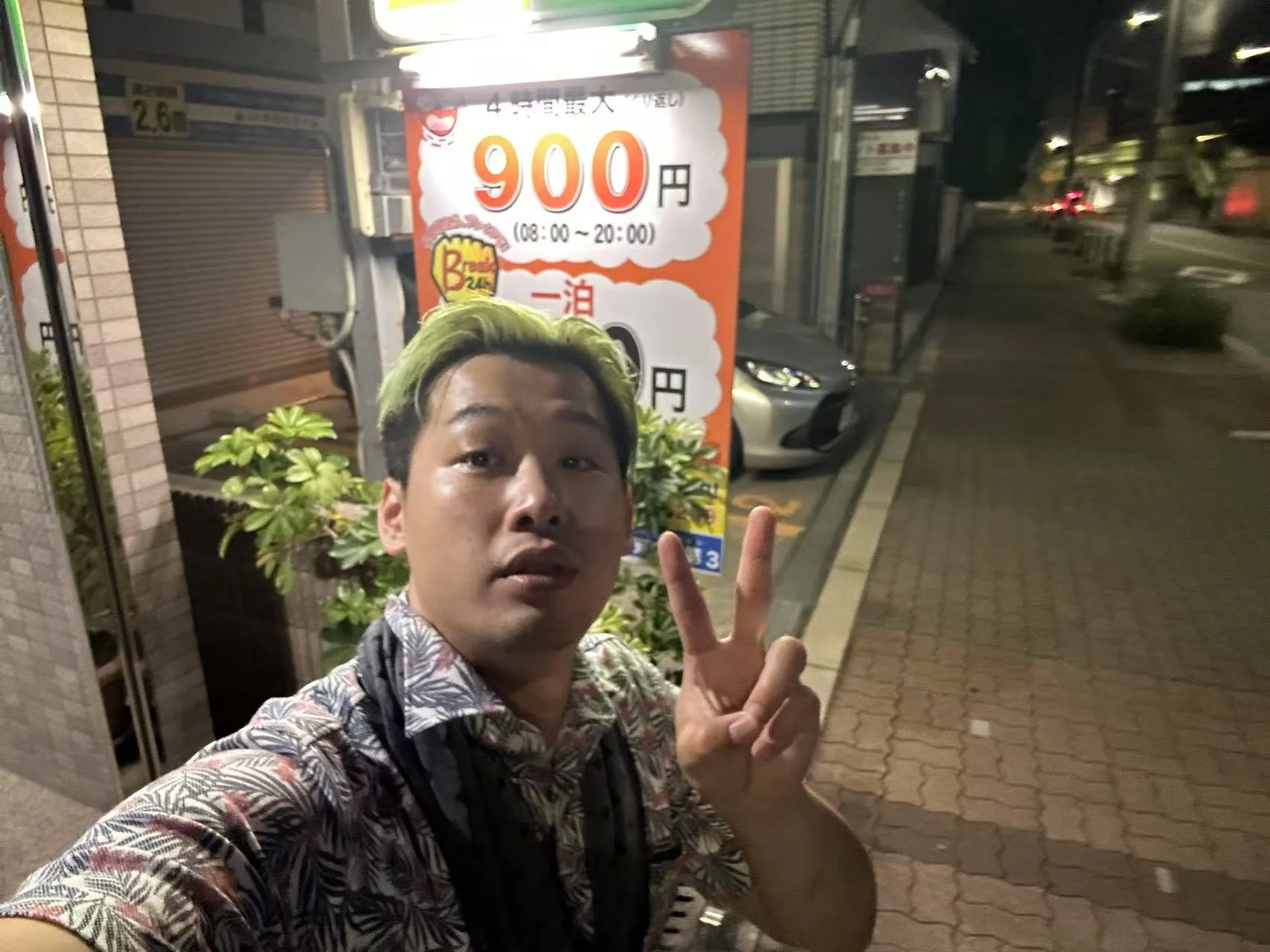 A young man taking a selfie on a city sidewalk at night, making a peace sign. Behind him, there is a sign advertising a price of 900 yen for a service available from 8:00 to 20:00, with some plants and a parked car nearby.