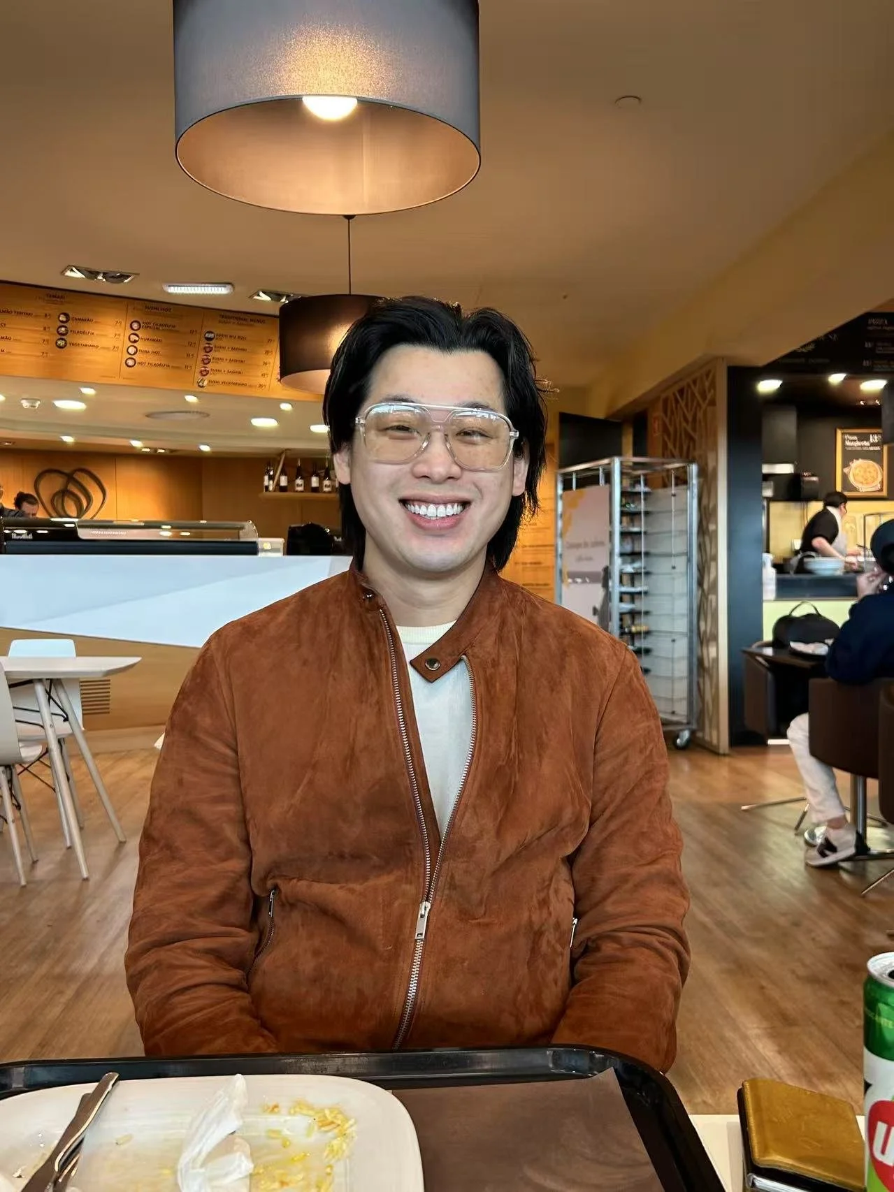 A man with black hair, glasses, and a brown suede jacket smiling at a restaurant. There is an empty plate in front of him, and the restaurant has a warm, modern interior with wood accents and bright lighting.