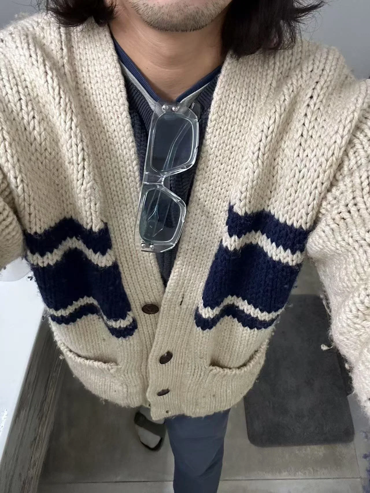 Person wearing a beige knitted cardigan with navy blue zigzag patterns, blue jeans, and sunglasses hanging on the cardigan's collar, standing in front of a mirror.