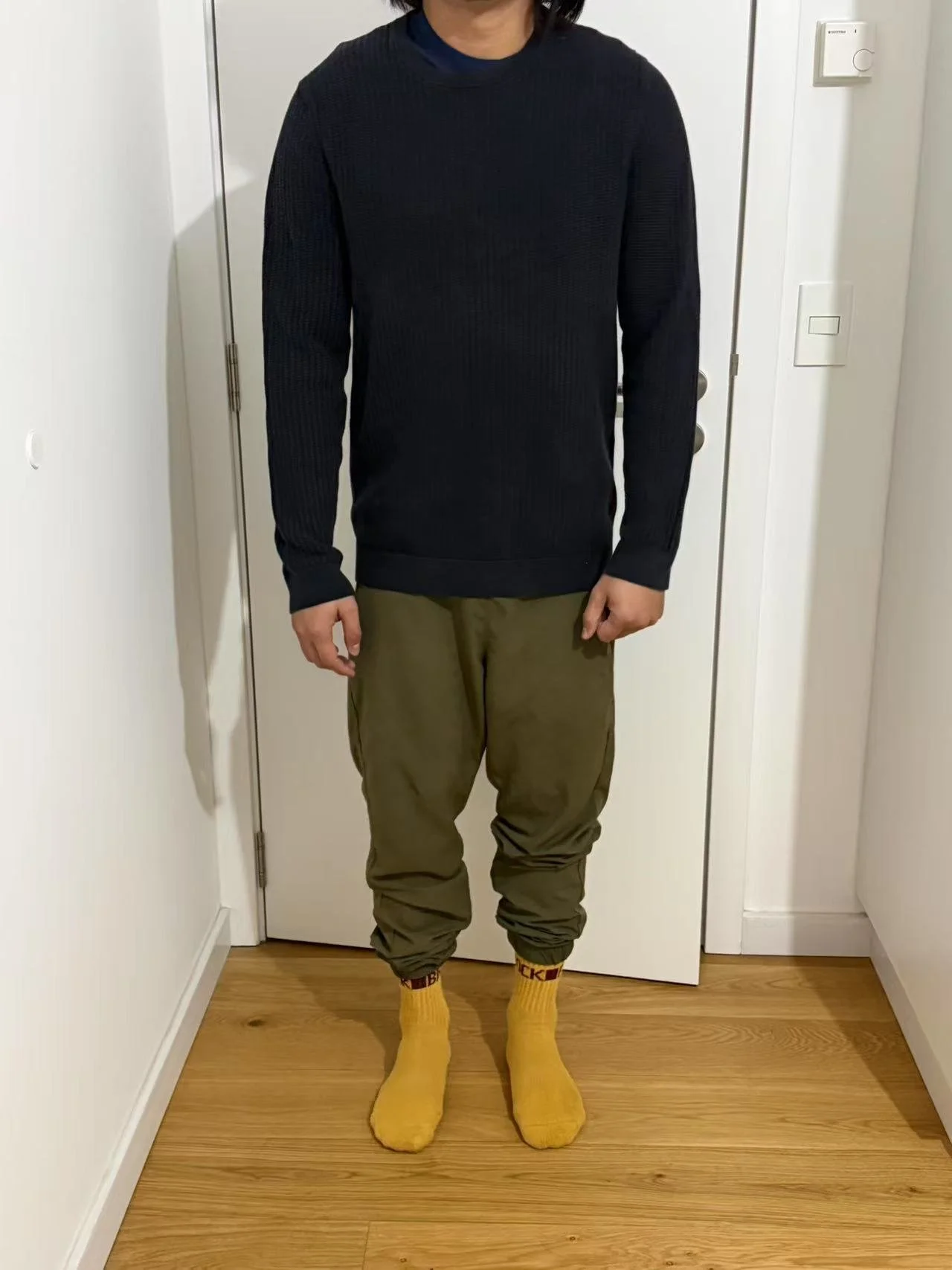 Person standing in a doorway wearing a black sweater, olive green pants, and yellow socks.