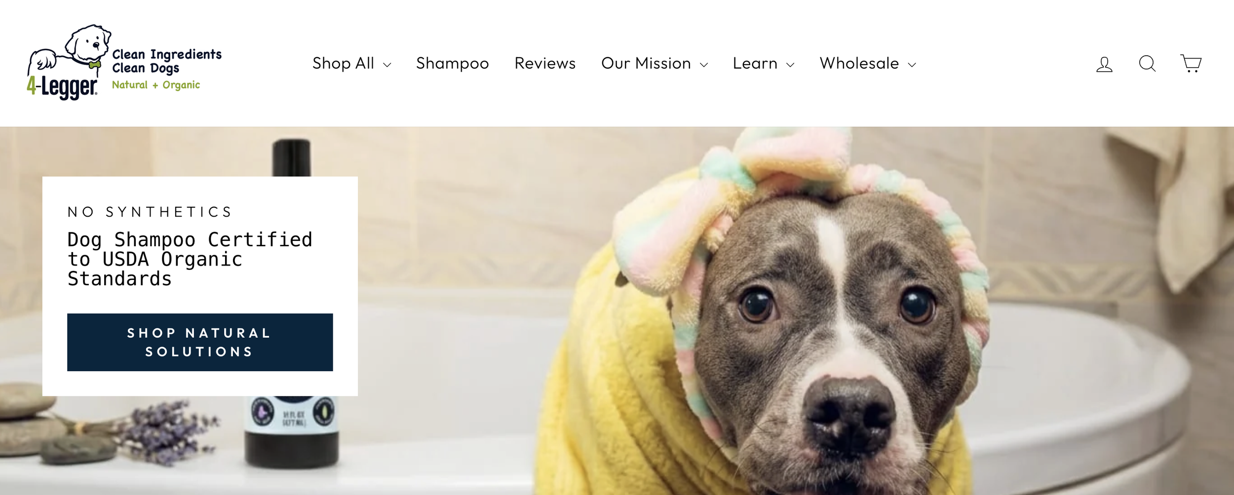A dog with a colorful towel on its head and wearing a yellow hoodie, standing in a bathroom with a beige tile background.