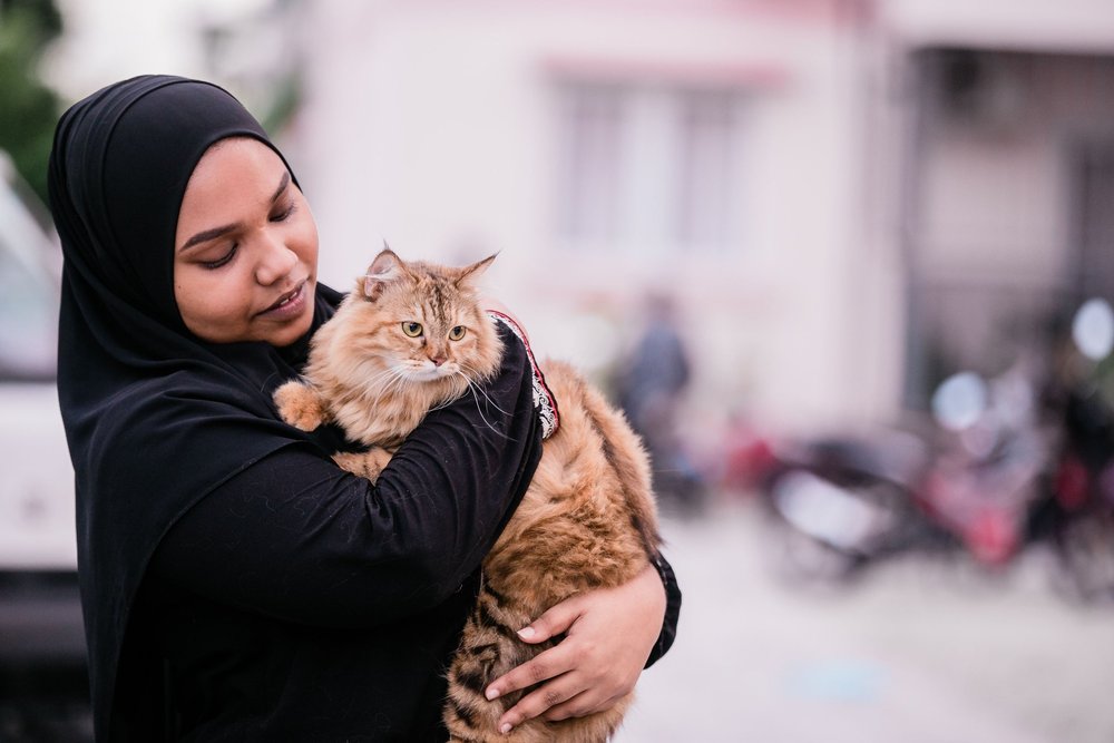 A person in a black headscarf cradling a long-haired ginger cat outdoors.