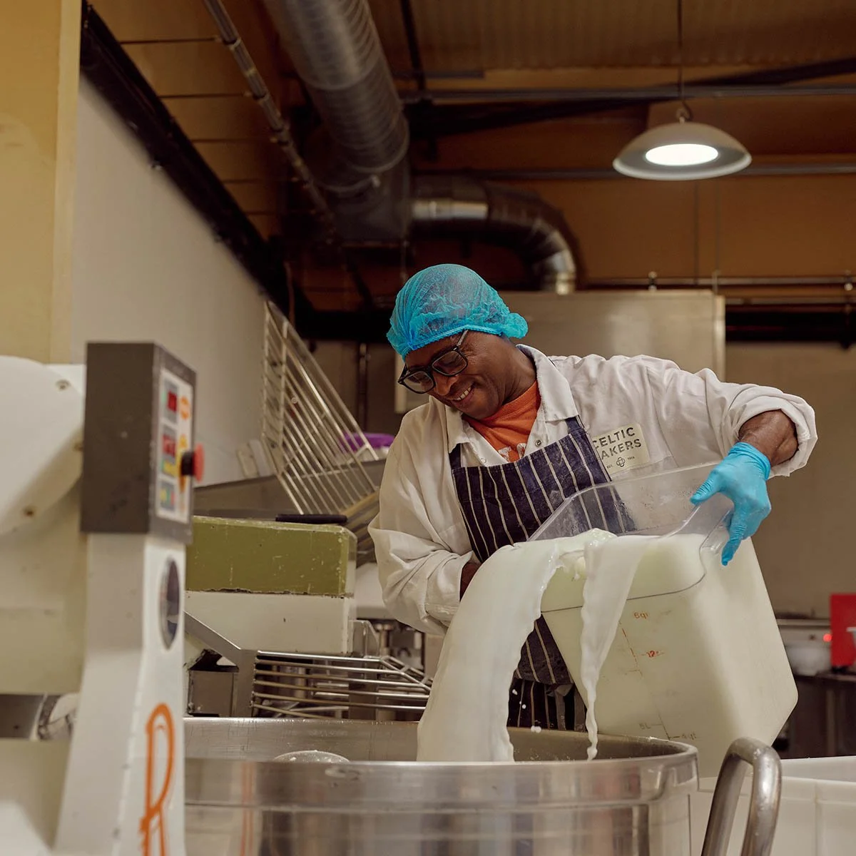 A man wearing a blue hair net, glasses, and a striped apron, smiling while pouring milk or cream from a large container into a metal vat in a dairy or cheese-making facility.