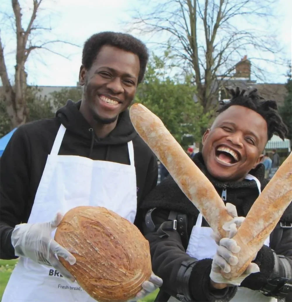 Two men smiling and holding loaves of bread and baguettes outdoors in a park with trees in the background.