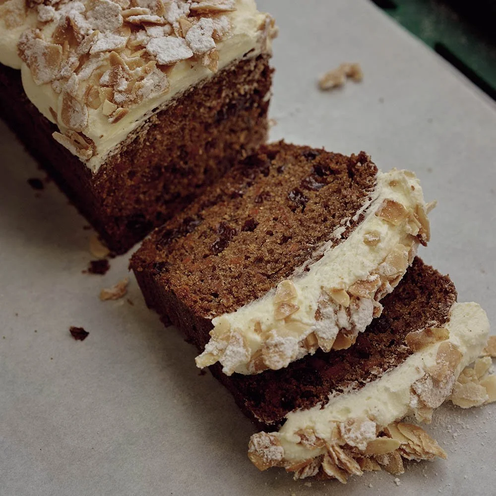 A sliced chocolate loaf cake with white frosting and chopped almonds on top, placed on parchment paper.