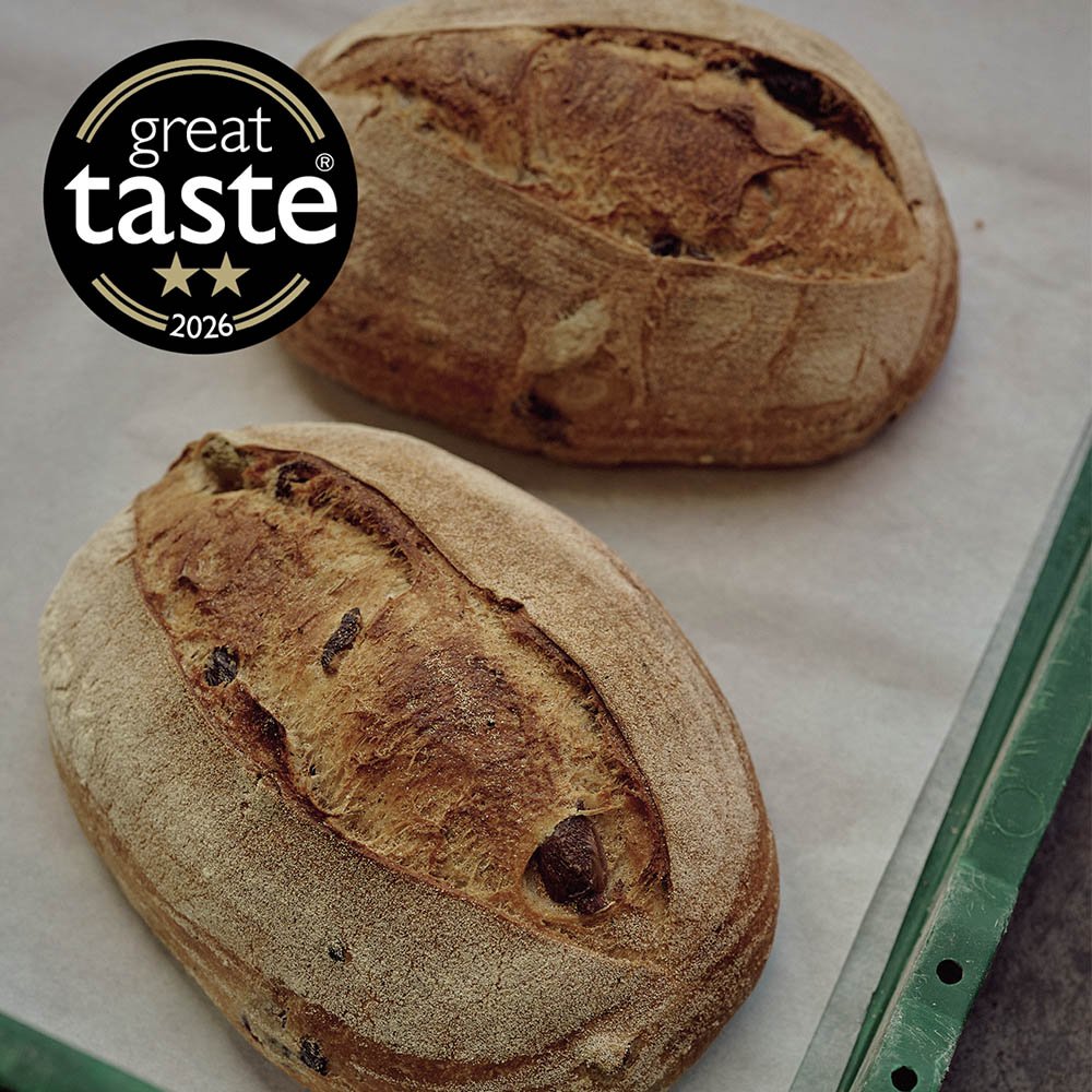 Two freshly baked oval-shaped artisan bread loaves with a rustic crust and scoring on top, resting on parchment paper. A 'Great Taste' award badge with two stars and the year 2026 is visible on the top left.