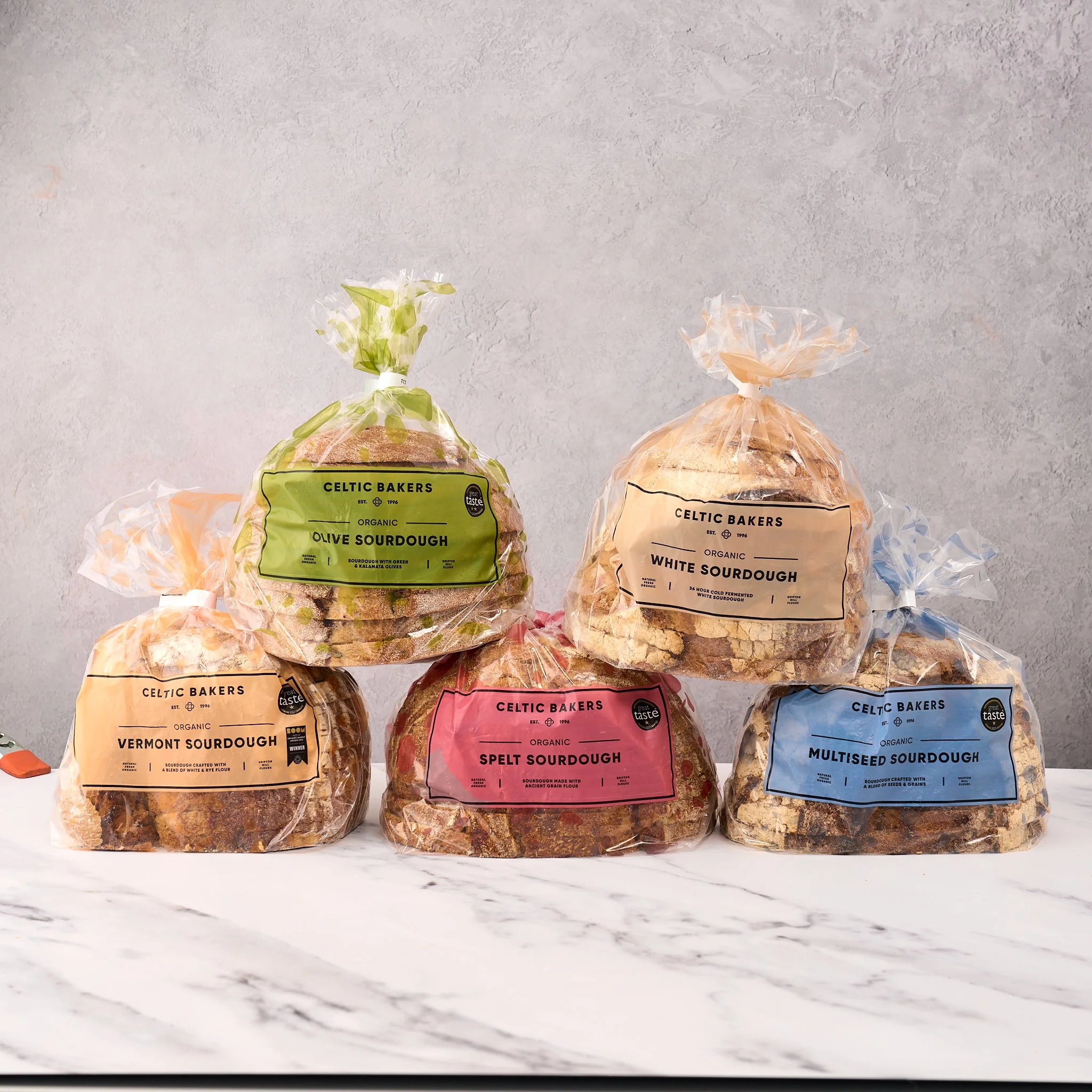 Three loaves of sourdough bread in colorful plastic bags on a marble surface, with a light gray textured wall in the background.