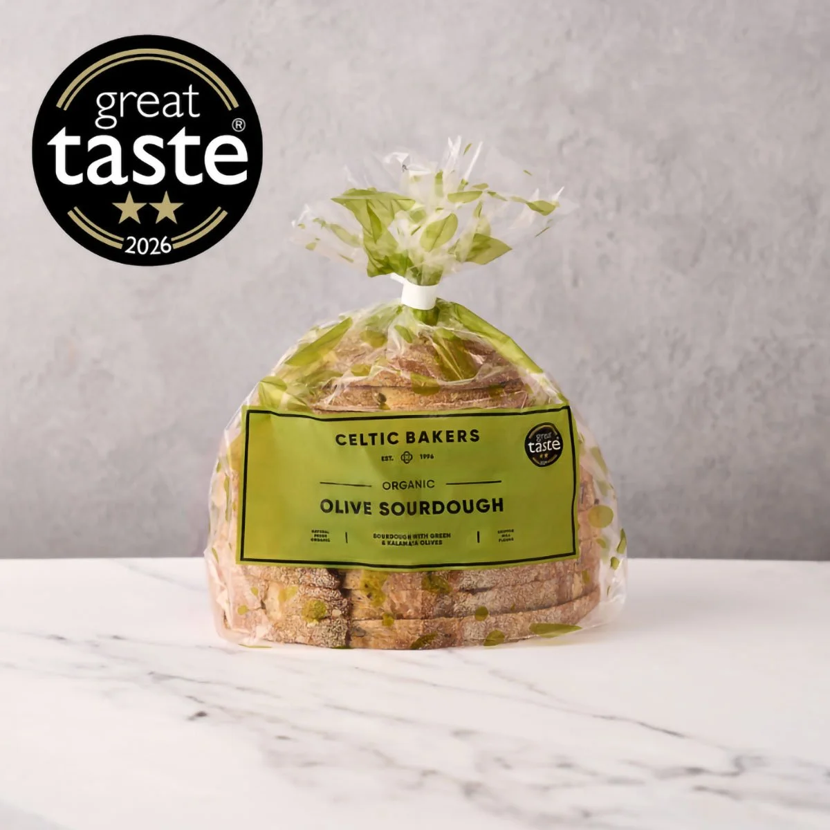 Package of Celtic Bakers organic olive sourdough bread in clear plastic bag with green label and black text, placed on a white marble surface with a gray wall background.