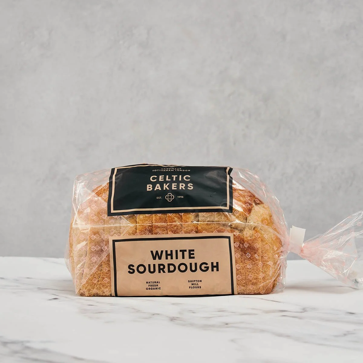A loaf of white sourdough bread in transparent packaging from Celtic Bakers, sitting on a white marble surface with a light gray background.