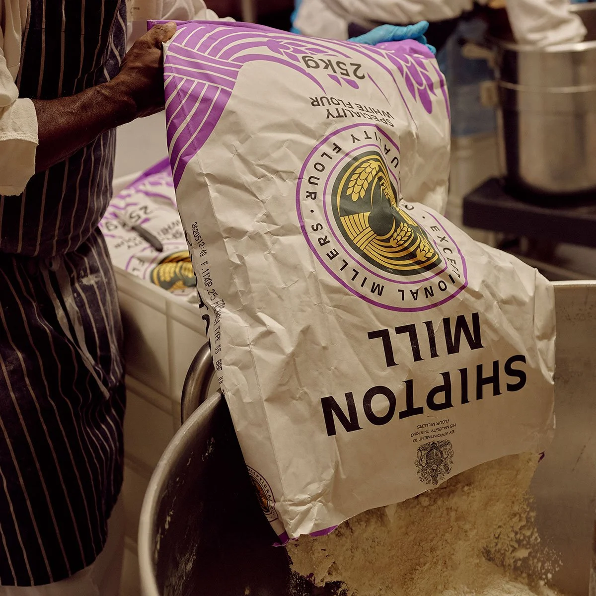 A crumpled bag of plain white flour with purple and black labeling being held by a person wearing a striped apron.
