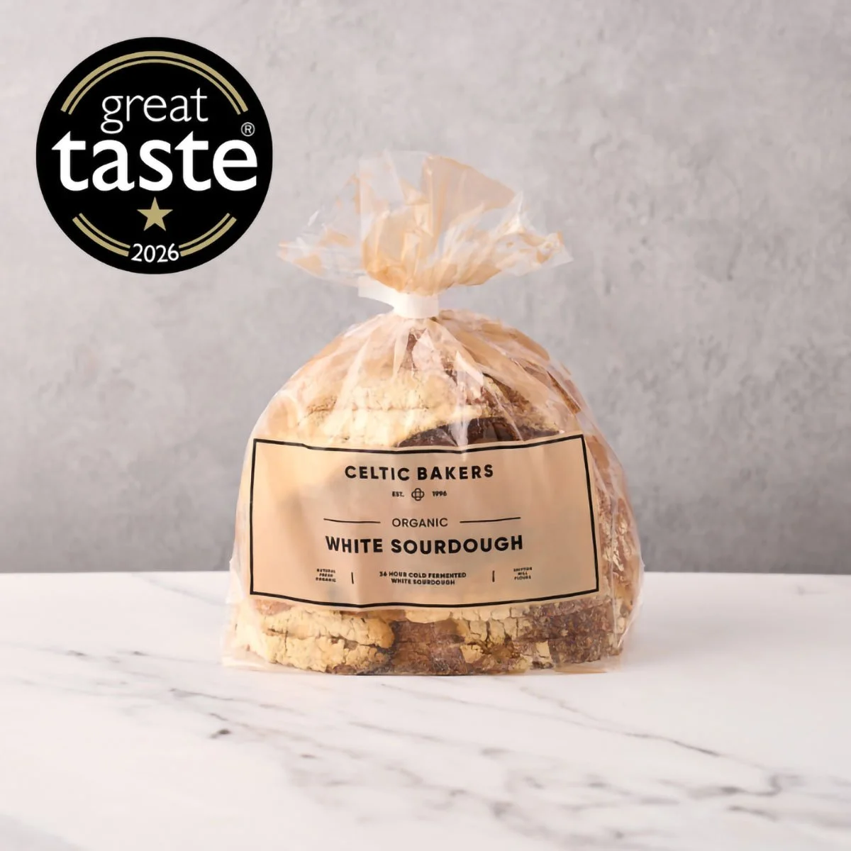 A clear plastic bag of organic white sourdough bread from Celtic Bakers on a white marble surface with a plain grey background, featuring a Great Taste 2026 award badge in the upper left corner.