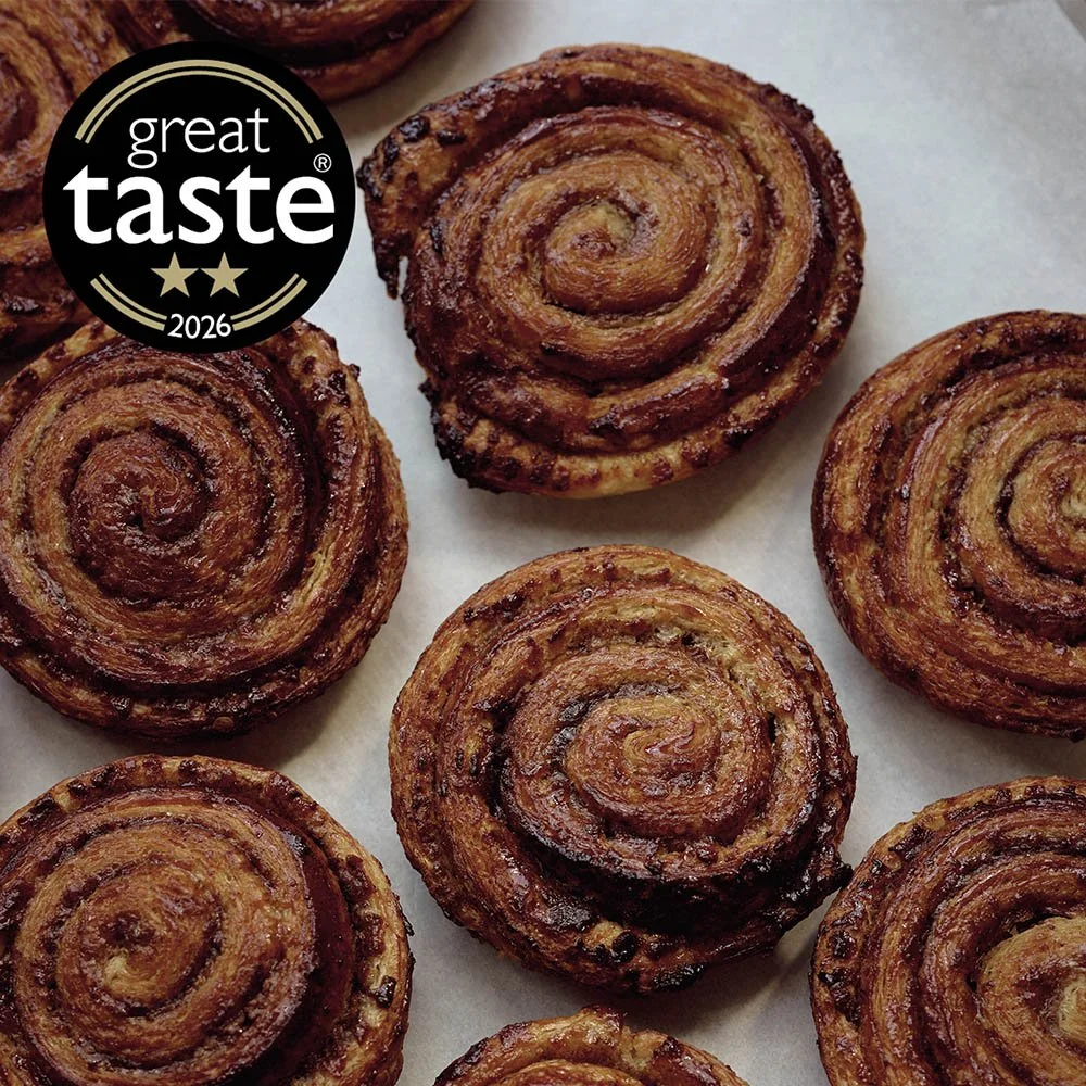 Close-up of several cinnamon rolls on parchment paper, with a Great Taste award badge from 2026 in the top left corner.