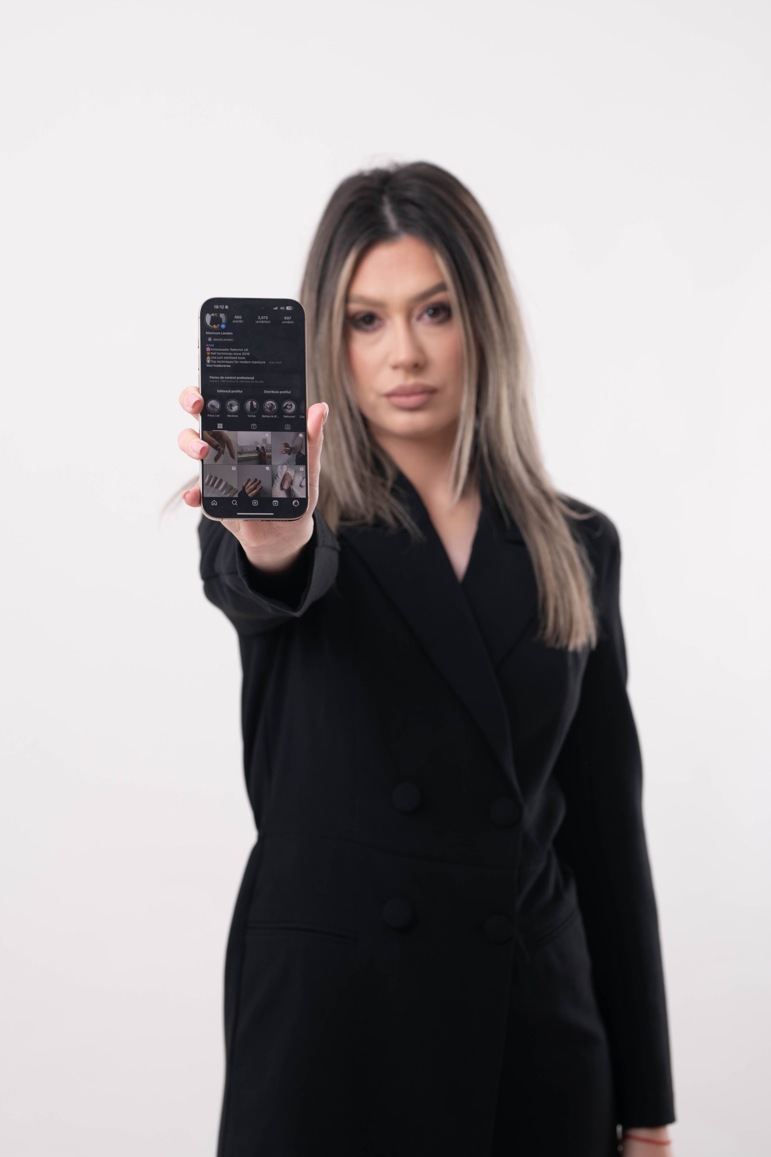 A woman with long hair wearing a black blazer, holding a smartphone towards the camera, with a plain white background.