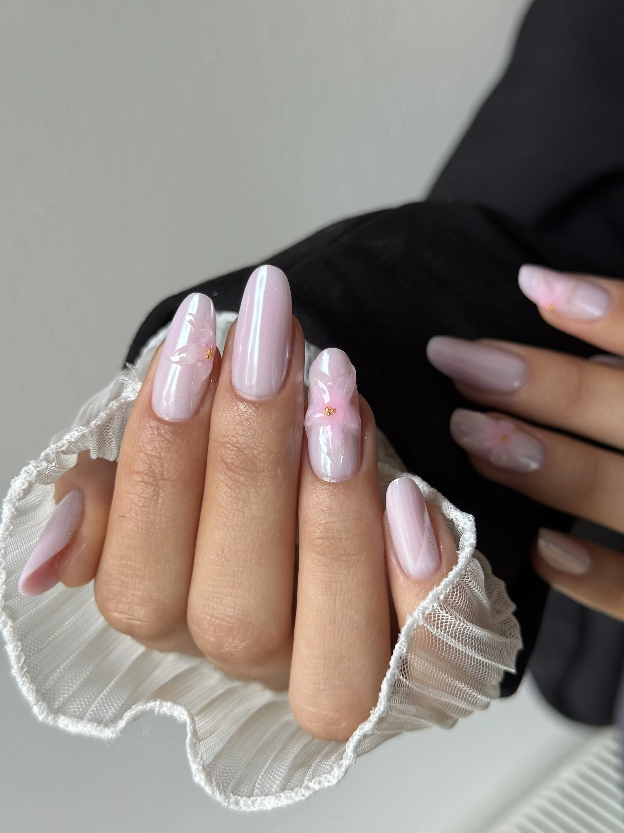 Close-up of a person's hand with long, oval-shaped nails painted in a light, shiny pink with small gold accents, held over a lace-trimmed fabric.
