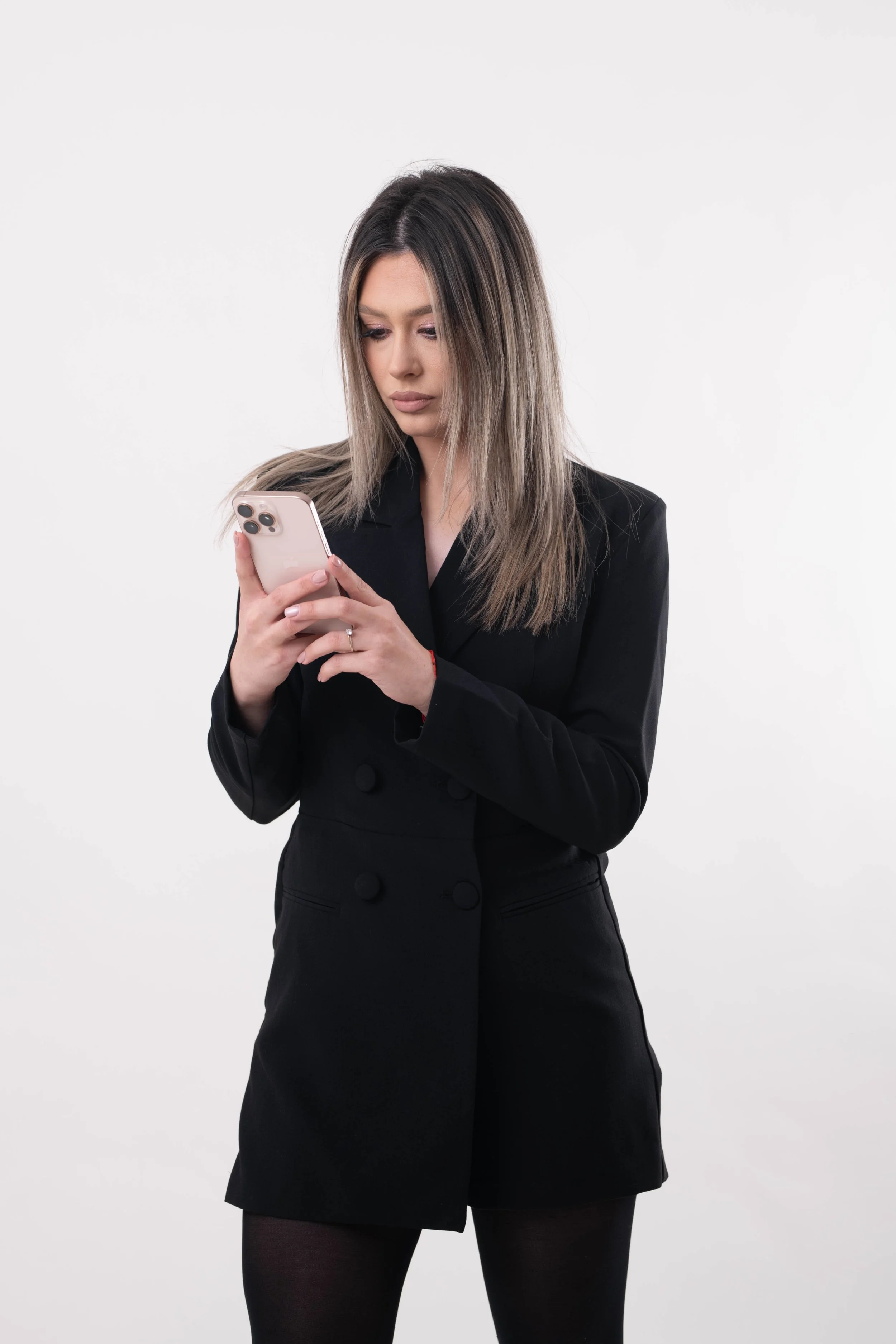 A woman with shoulder-length blonde hair looking at her smartphone against a plain white background. She is wearing a black blazer and skirt, with black tights.