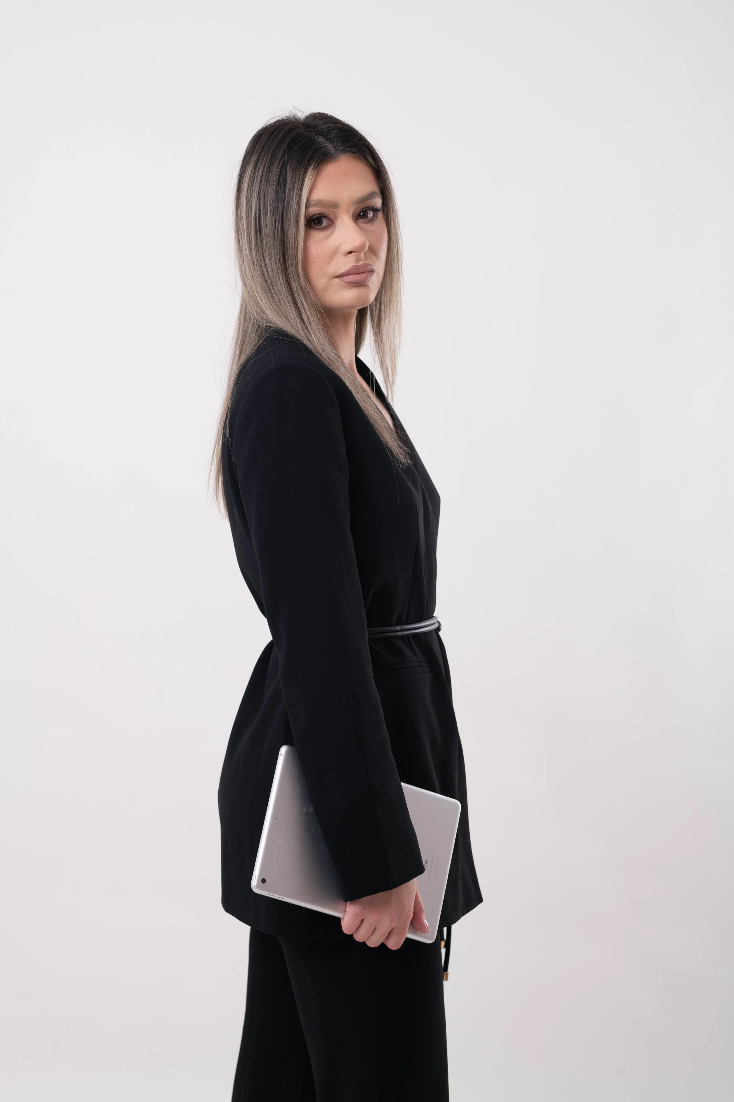 A woman with straight, ombré hair standing in profile, dressed in a black suit, holding a silver laptop, against a plain white background.