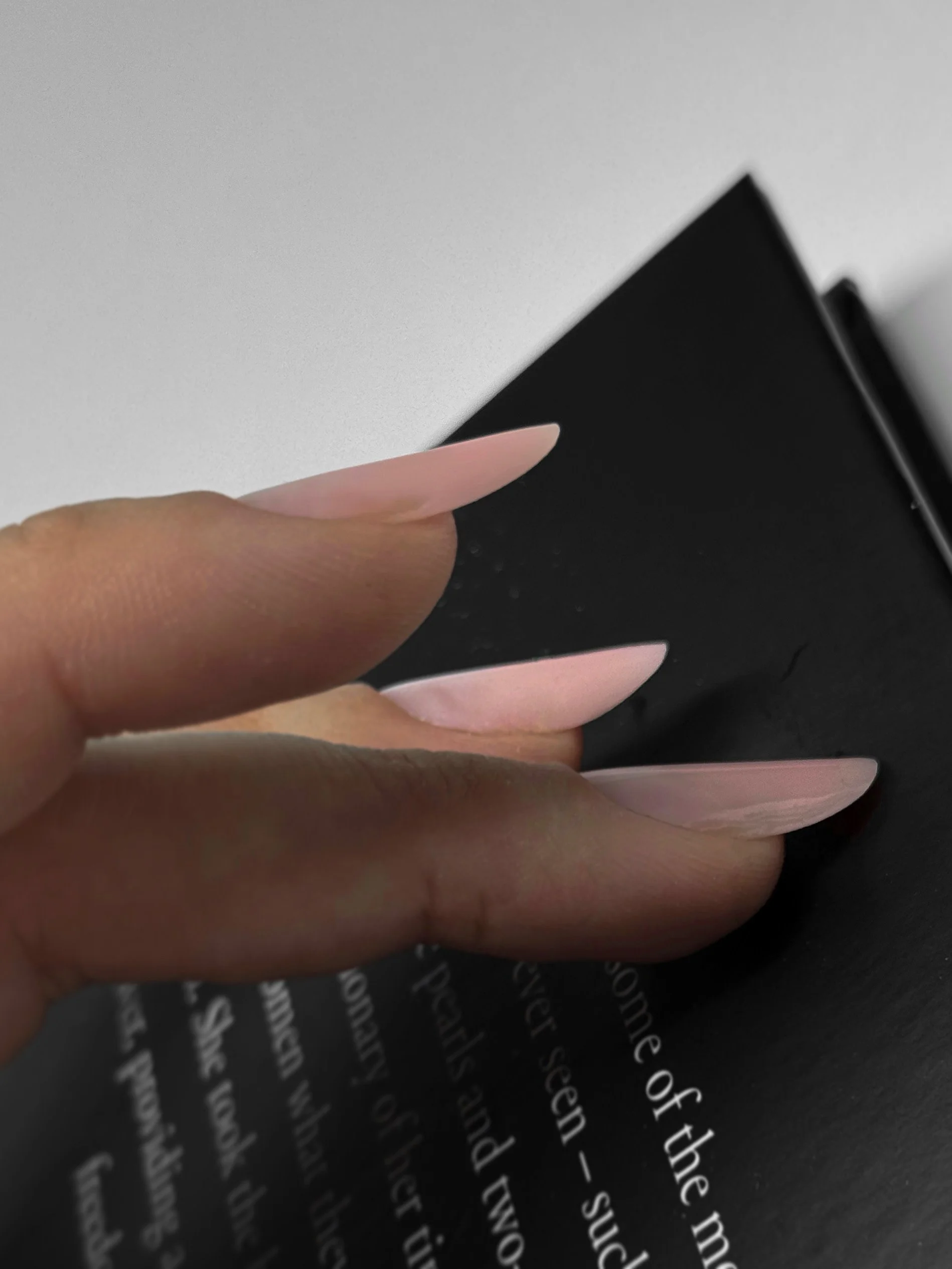 Close-up of a person's hand with long manicured nails holding a black book or journal against a white wall and a black surface with text.