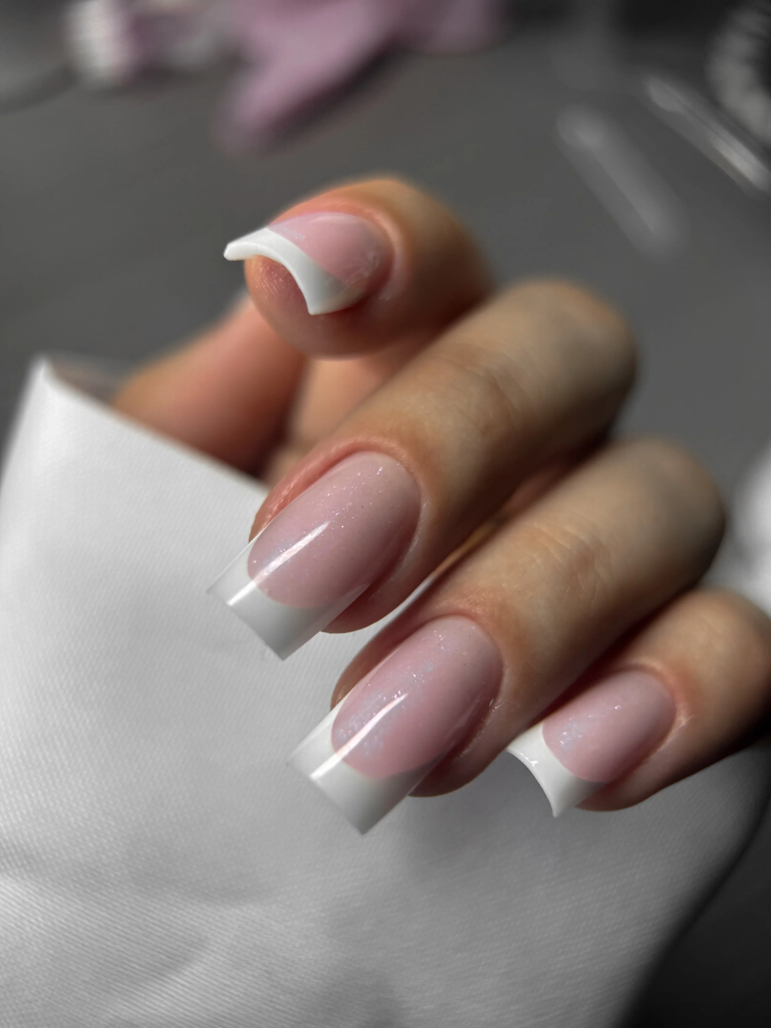 Close-up of a person holding a nail tip with a natural-looking French manicure nail extension on their fingers, resting on a white fabric surface.