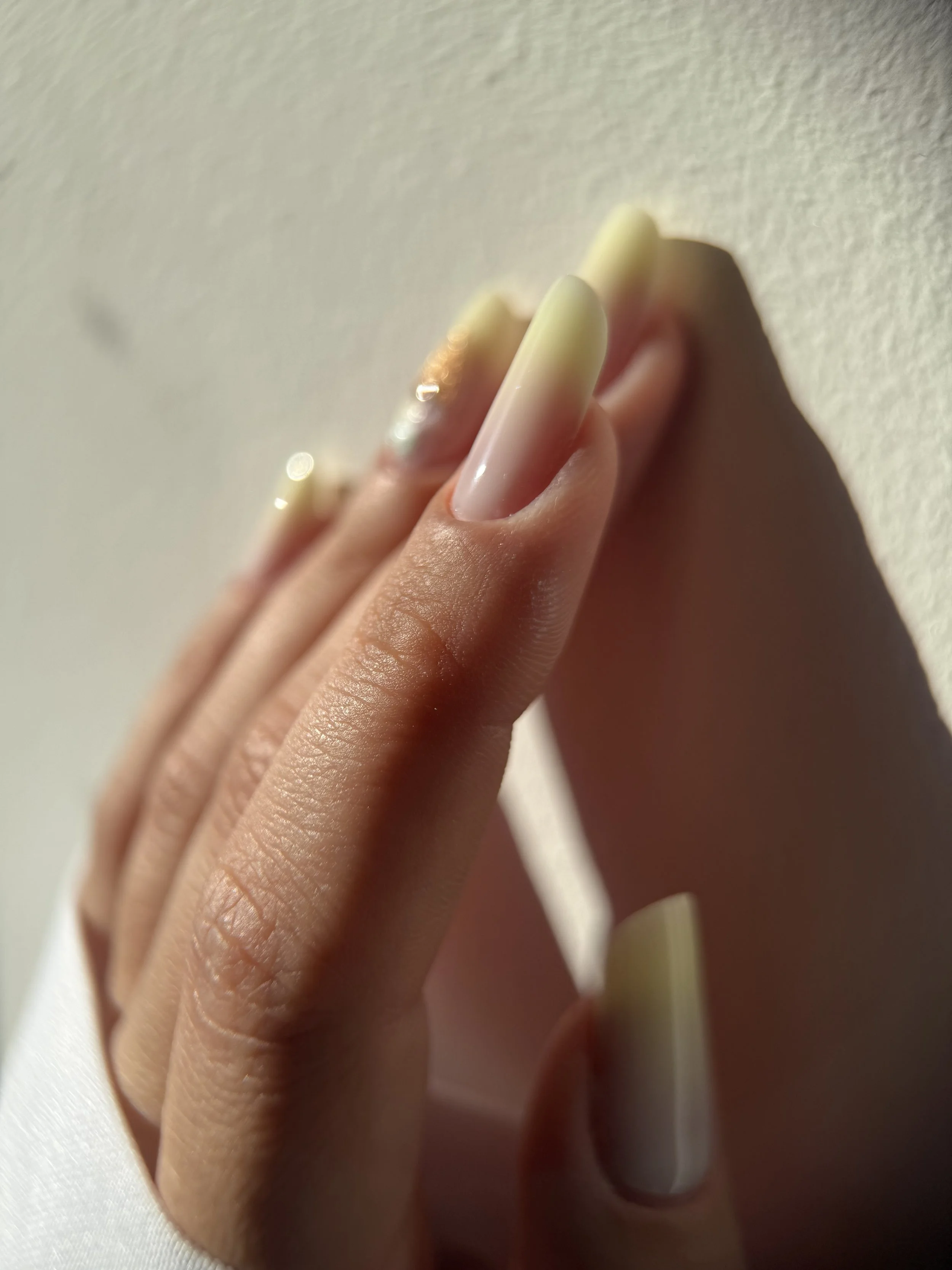 Close-up of a hand with long, square-shaped, pale yellow and pink ombre acrylic nails against a light wall.