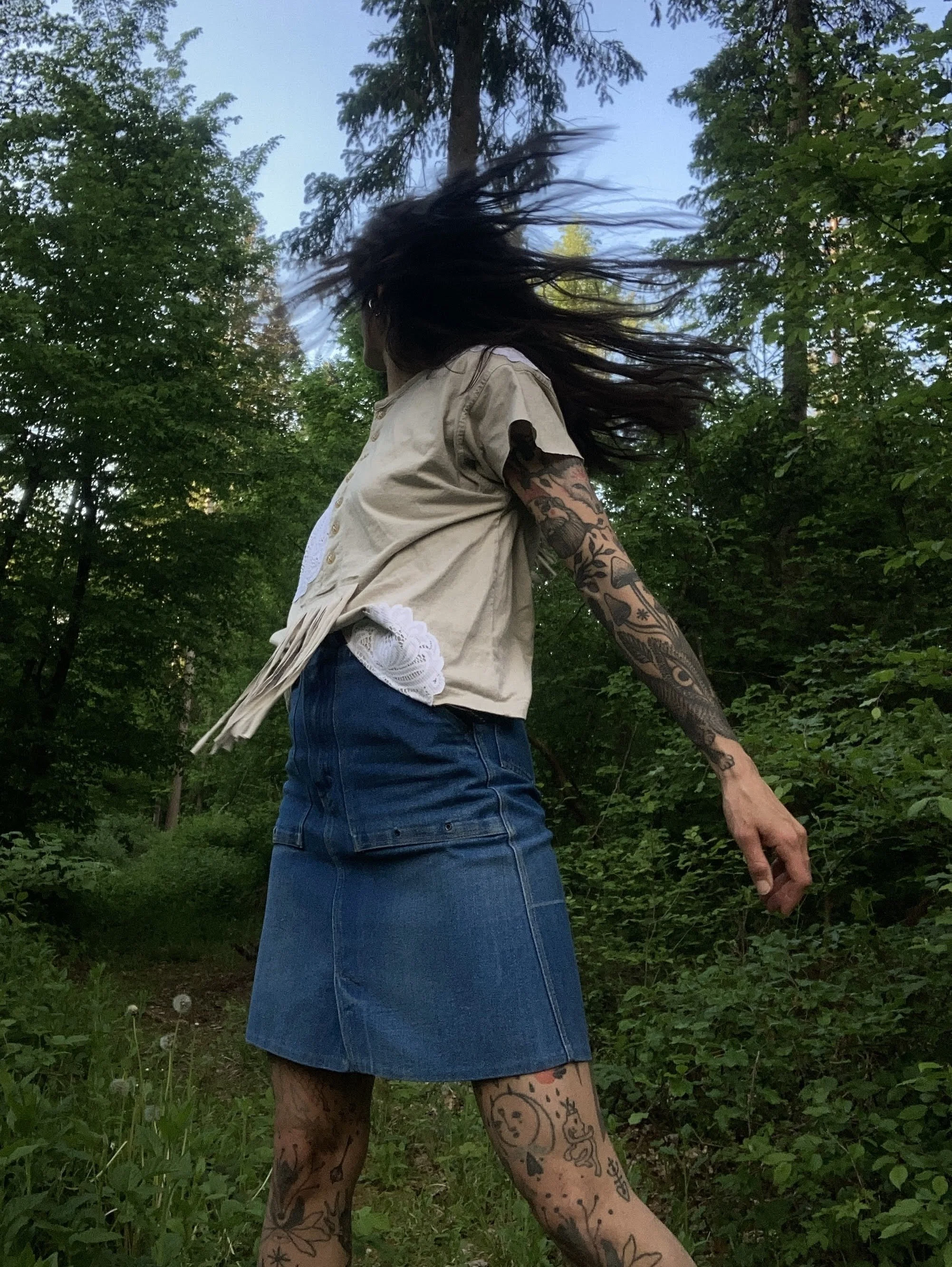 Person with long dark hair blowing in the wind, wearing a beige short-sleeved shirt and a denim skirt, standing outdoors among green trees with blue sky visible above.