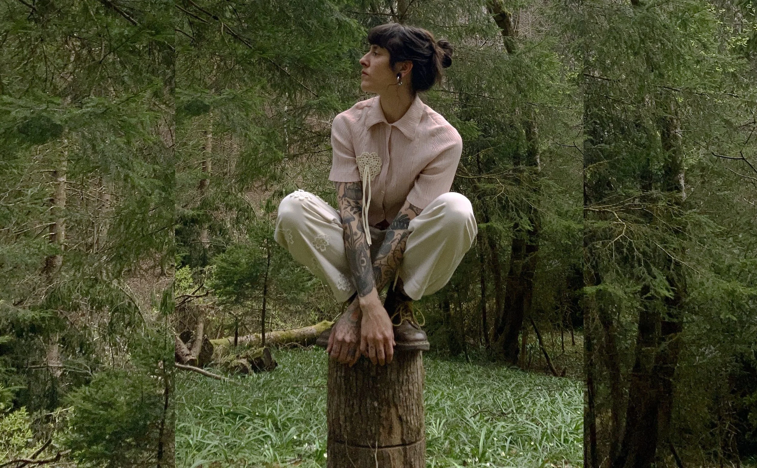 A woman with short dark hair, tattoos on her arms, wearing a beige shirt and pants, sits cross-legged on a tree stump in a lush green forest.