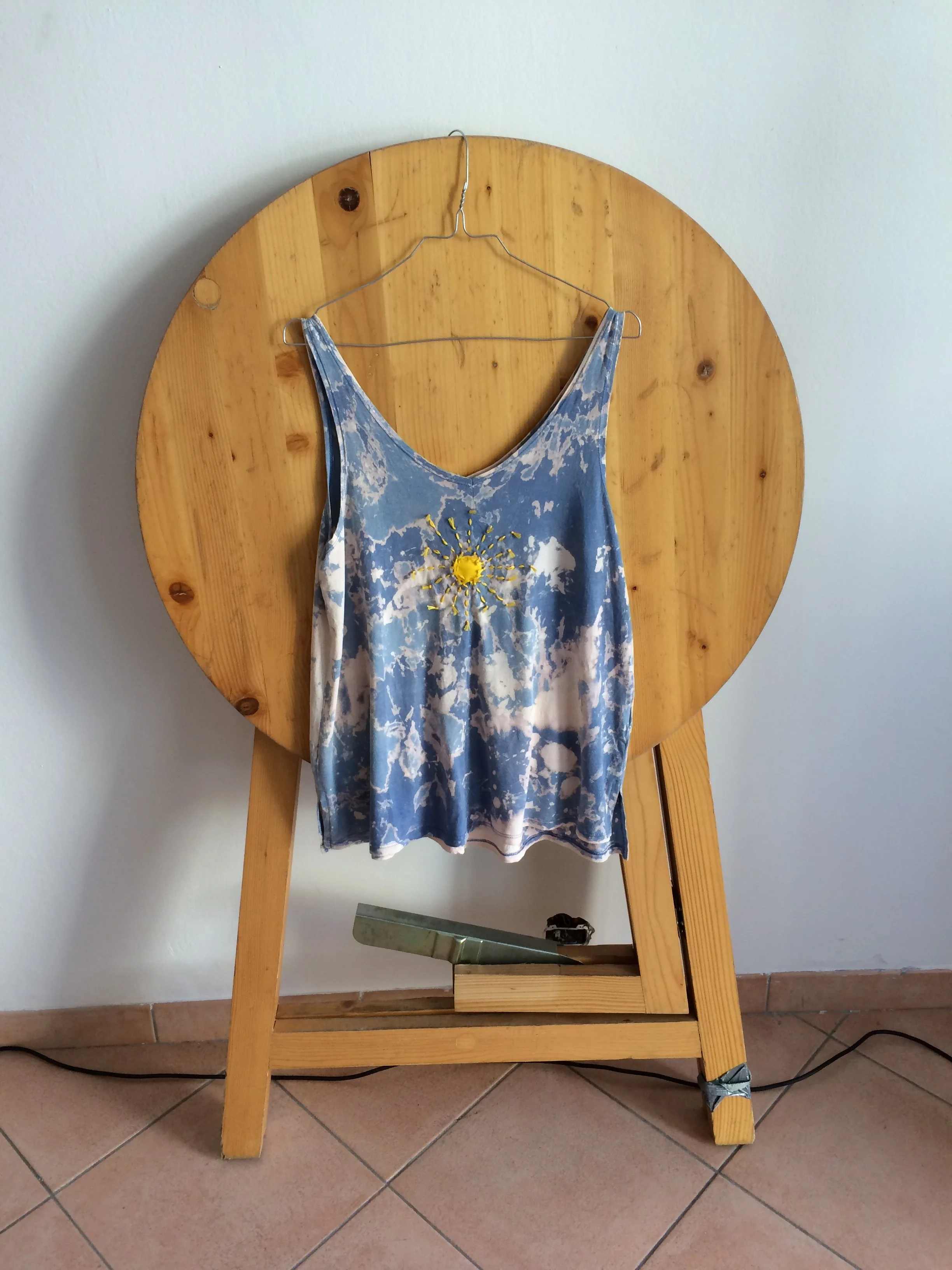 A wooden chair with a tie-dye dress hanging on a wire hanger, featuring a yellow flower design in the center.