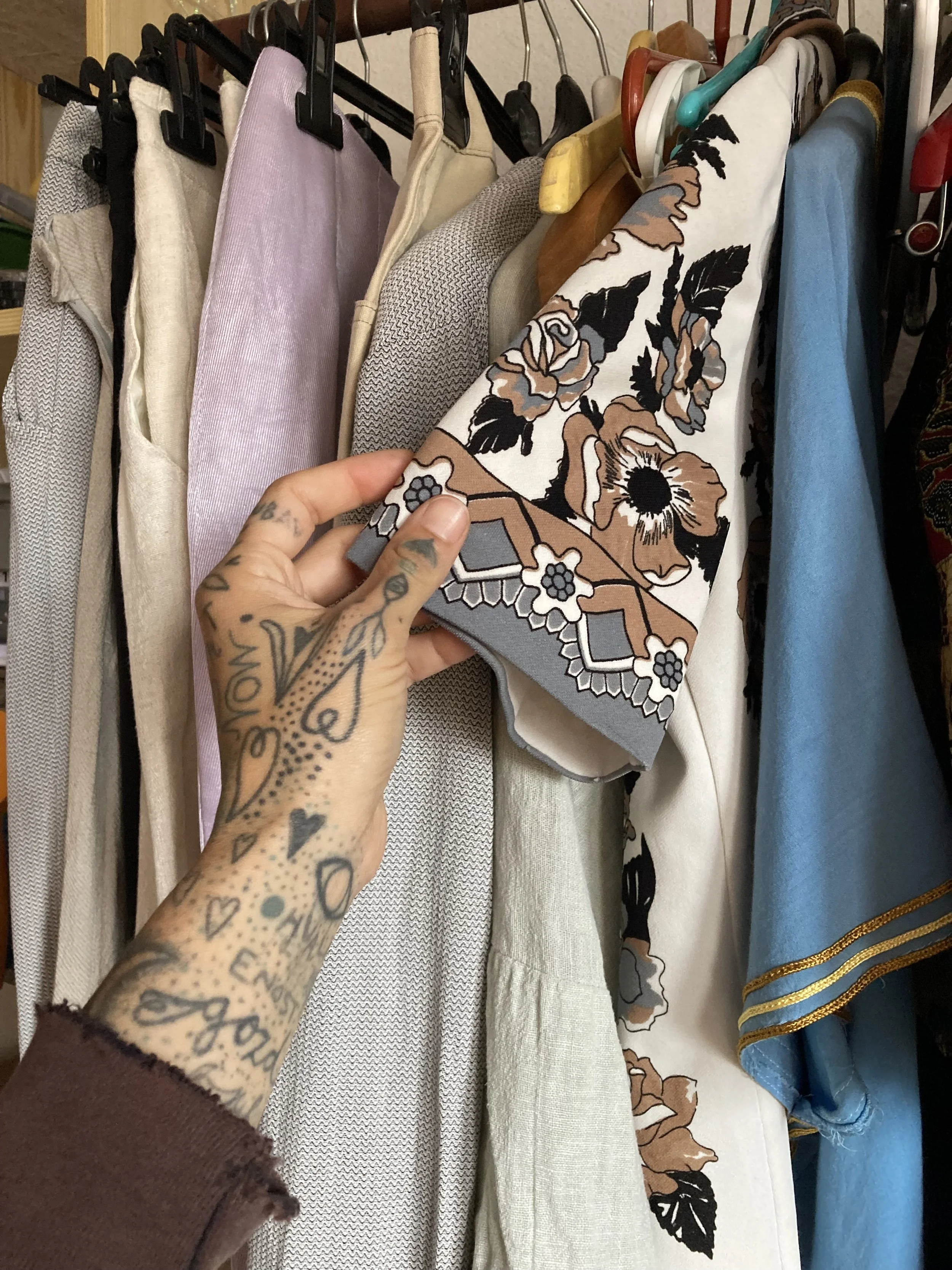 Close-up of a tattooed hand holding a floral patterned fabric among other garments hanging on a clothing rack.