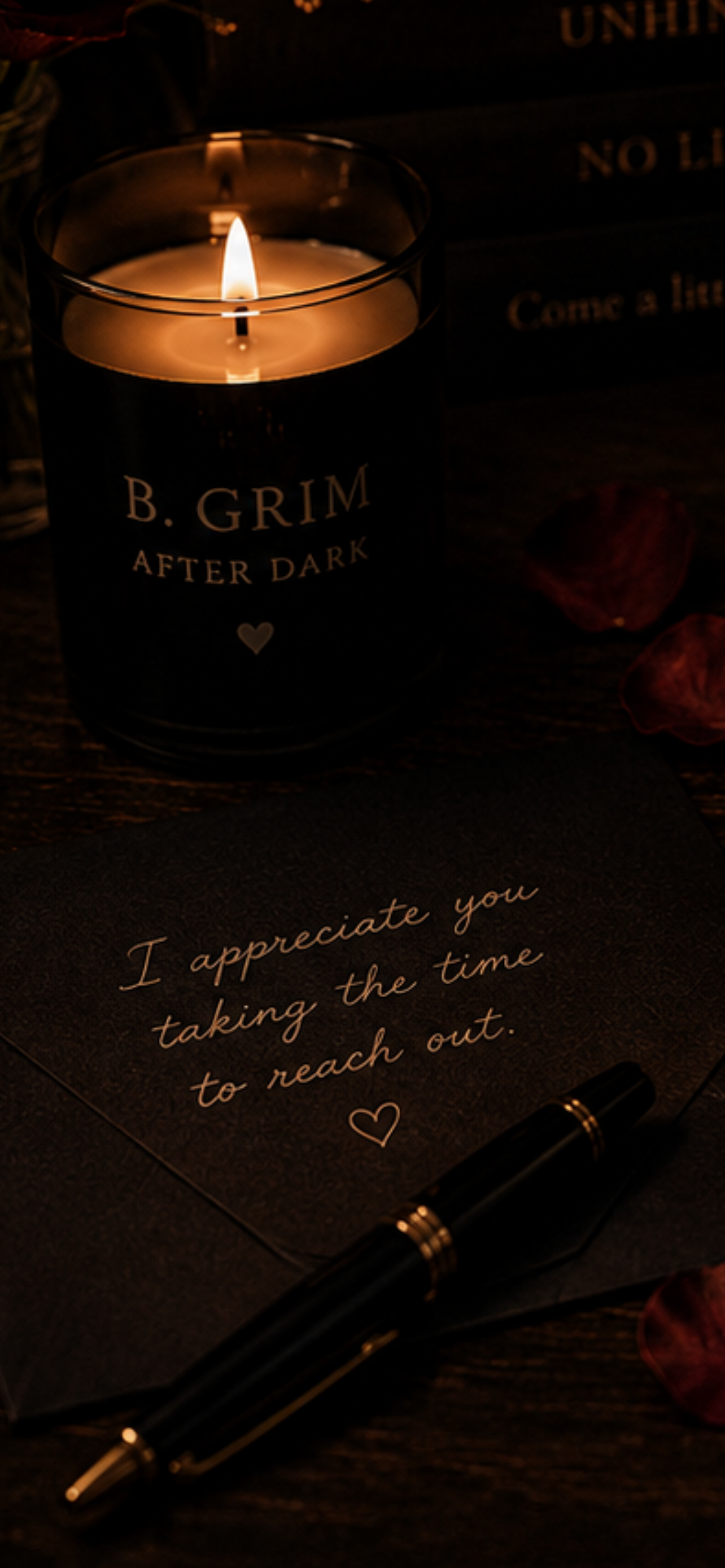 A lit candle in a dark glass holder with the label 'B. Grimm After Dark' on it, a handwritten note that says 'I appreciate you taking the time to reach out' with a small heart, a black pen with gold accents, and some flower petals on a wooden surface.