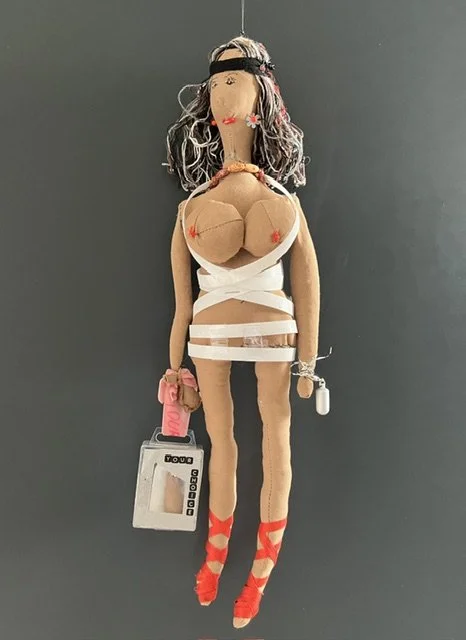 A handmade fabric doll resembling a woman, with gray and black hair, wearing a white outfit and red shoes, holding a small pink purse and a gray box.