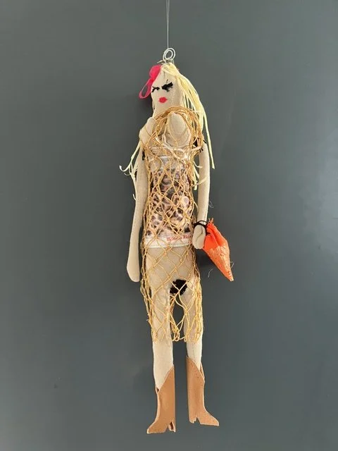 A decorative handmade doll hanging on a gray wall, dressed in a mesh-like outfit with a pink hair accessory and holding an orange clutch.