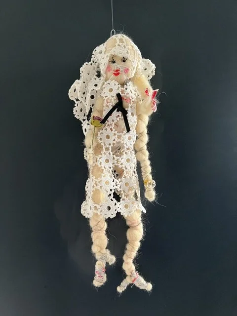 Doll made of lace doilies with braided hair and red lips, hanging against a dark background.