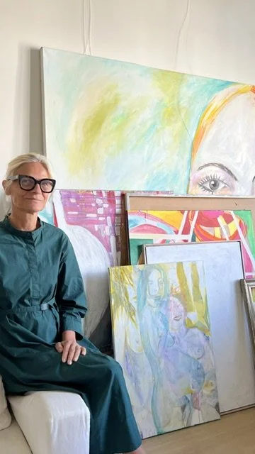 A woman with gray hair and glasses sitting on a white couch, surrounded by colorful abstract and portrait paintings leaning against the wall.