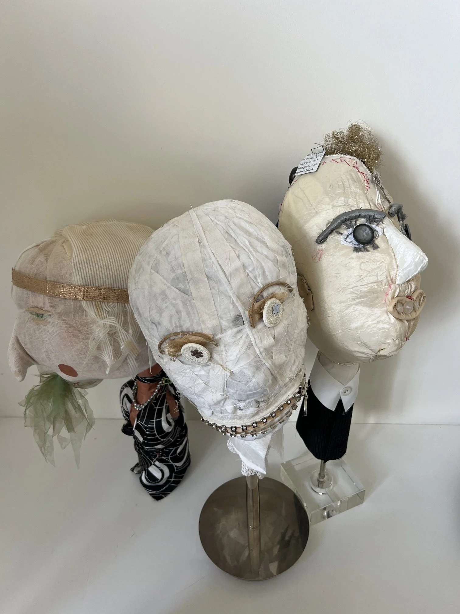 Three mannequin heads with artistic masks and headpieces, displayed on a metallic round stand against a plain white background.