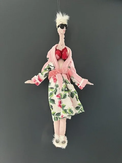 A doll with white hair, sunglasses, pink clothes, and fluffy footwear hanging against a dark background.