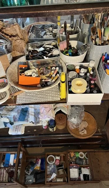 Cluttered desk with various art supplies, including paint bottles, brushes, markers, and paper, organized in containers and on surface.