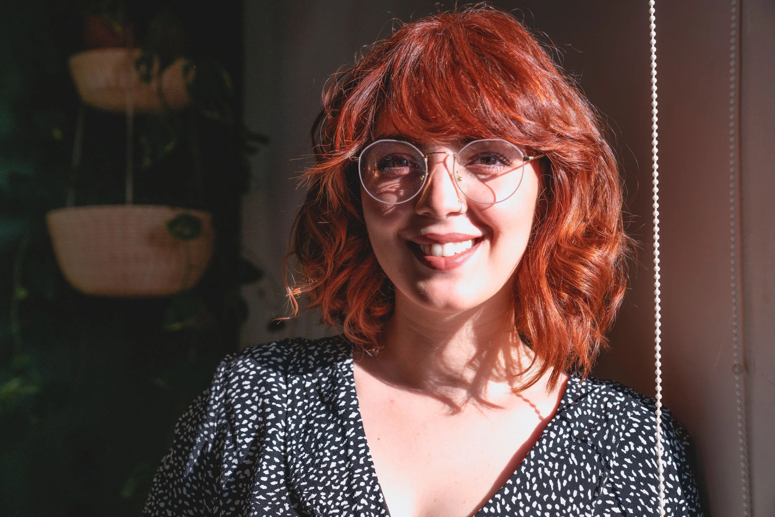 A young woman with curly red hair and glasses smiling, wearing a black and white dotted top, standing near a window with sunlight shining on her face.