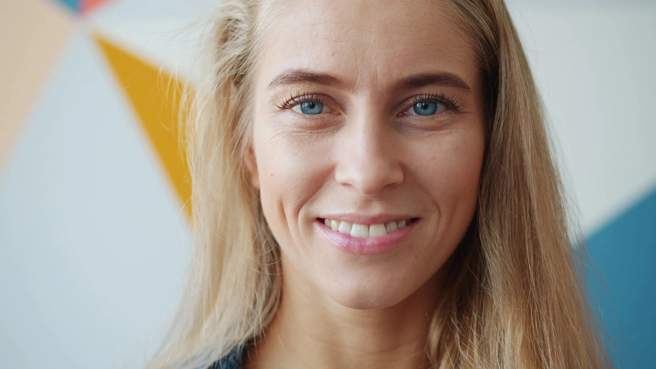 Close-up of a smiling woman with blonde hair and blue eyes, standing against a colorful background.