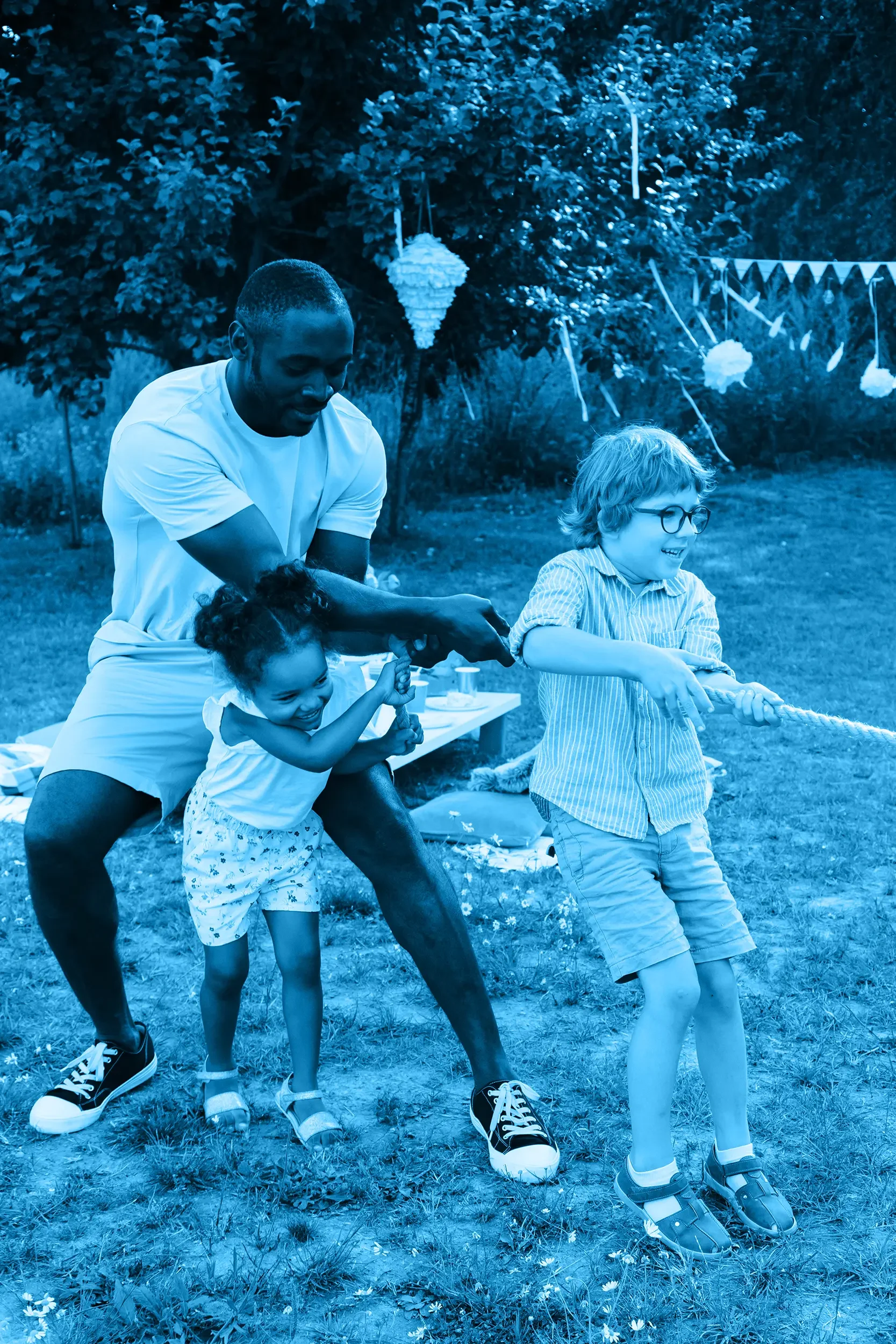 Group of people playing tug-of-war outdoors during daytime, with a father and two children pulling on a rope, surrounded by greenery and decorative lanterns hanging from trees.