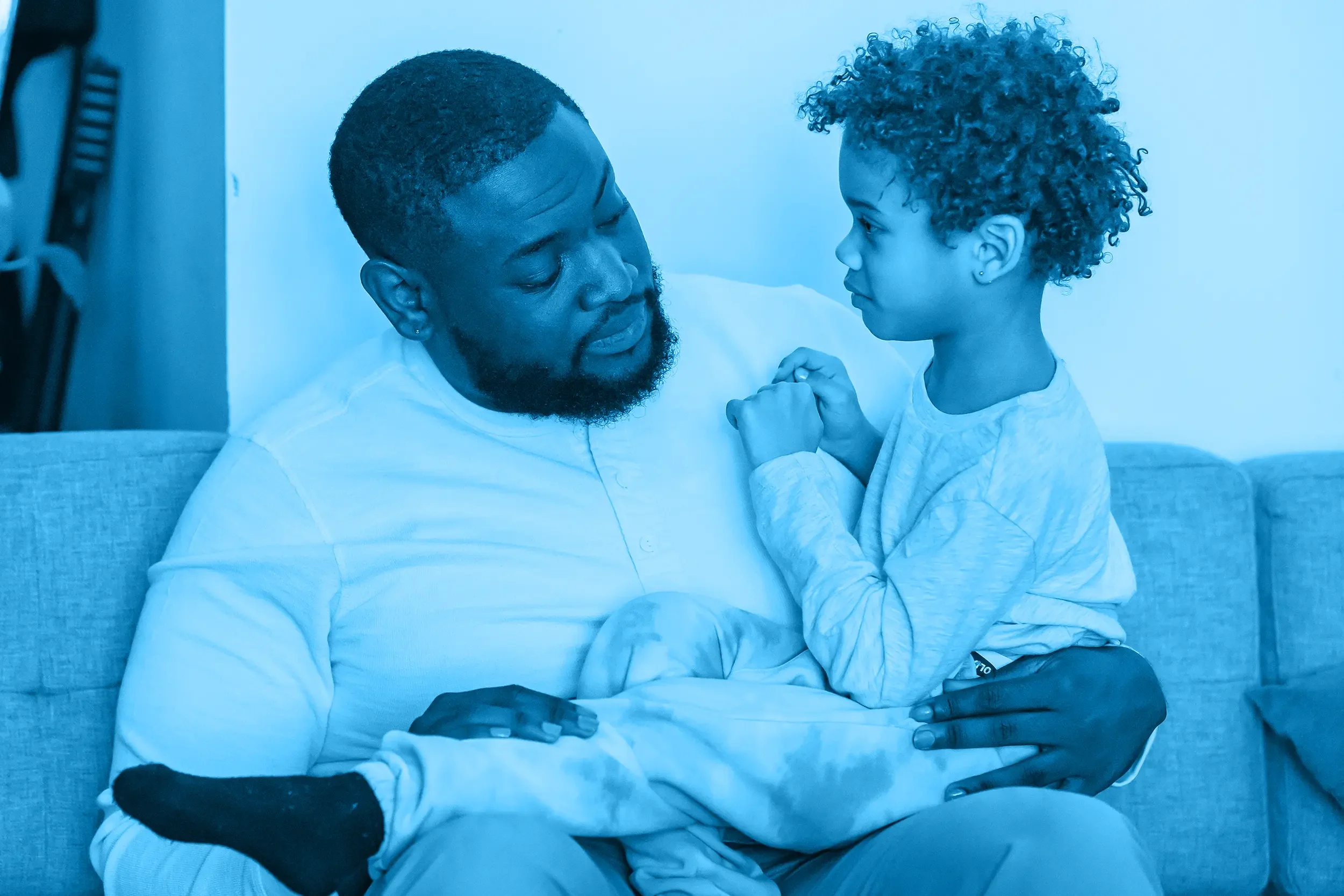 A father and a young girl sitting closely together, facing each other, with the dad’s eyes closed and the girl holding his hands in a gentle, emotional moment.