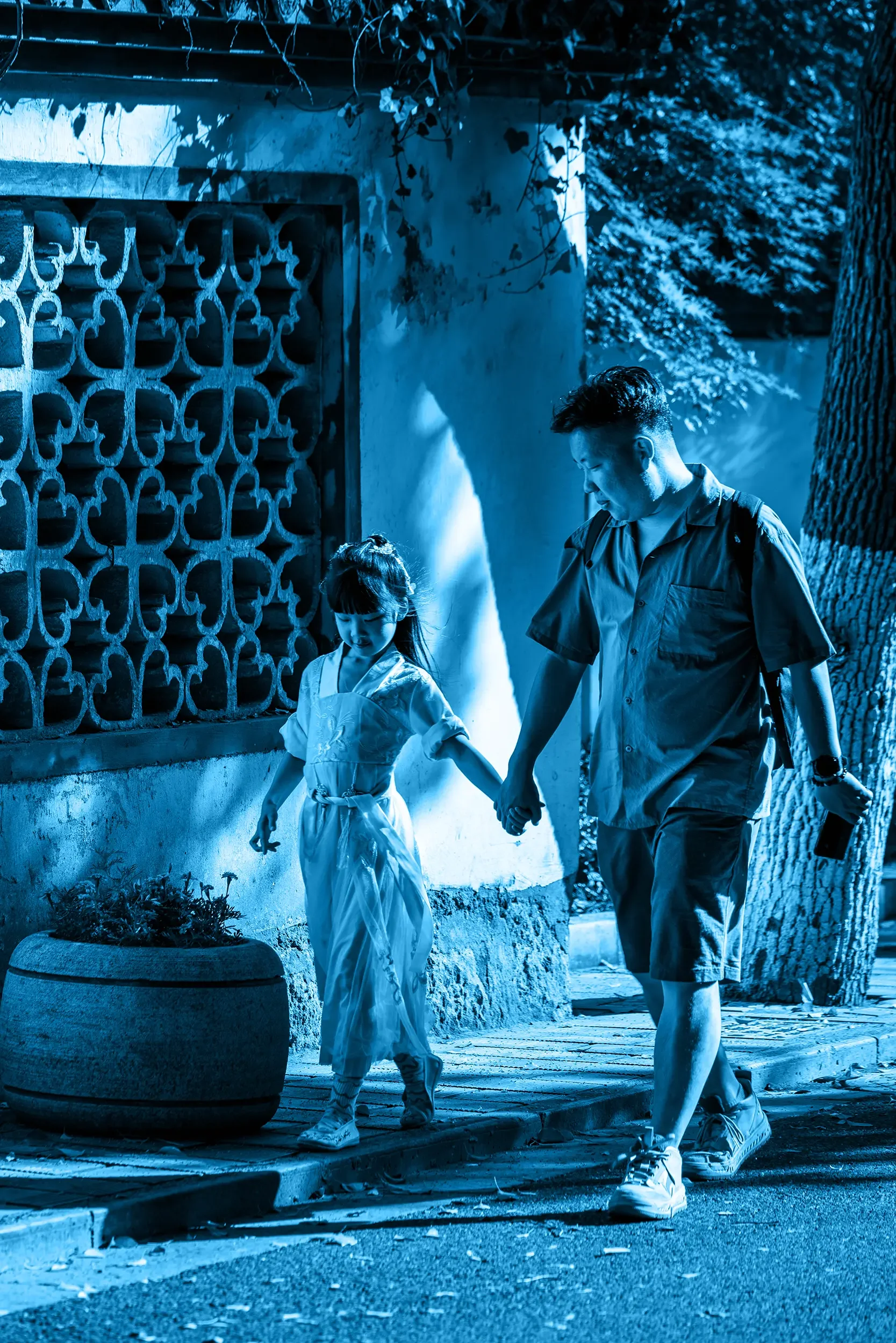 A  and a young girl holding hands while walking on a sidewalk at night, with a plant in a large round container nearby, tree trunks, a wall with patterned ventilation blocks, and a leafy tree in the background, all illuminated by blue-tinted lighting.