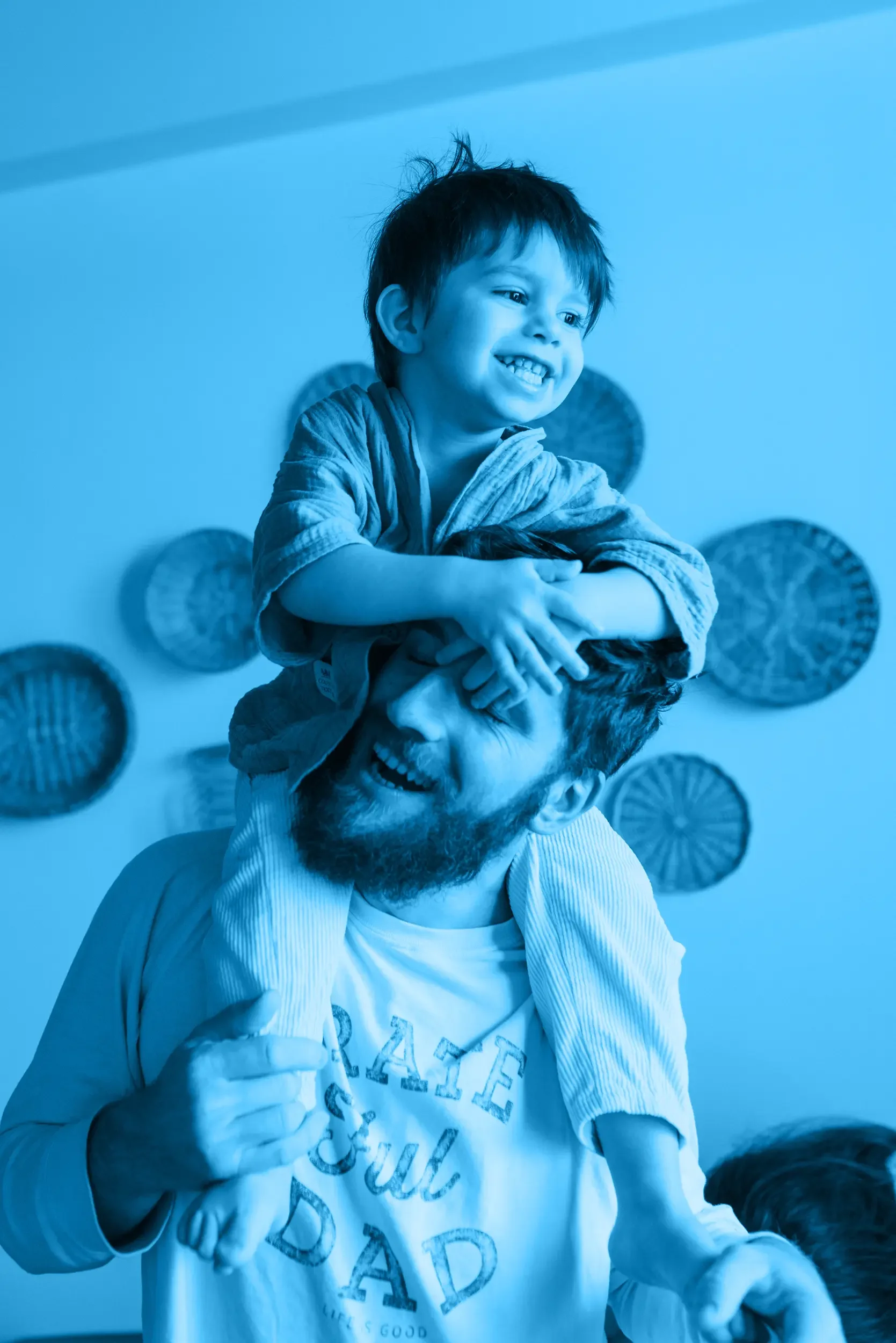 A young boy with dark, messy hair and a big smile, sitting on a father's shoulders, with their hands on his head.