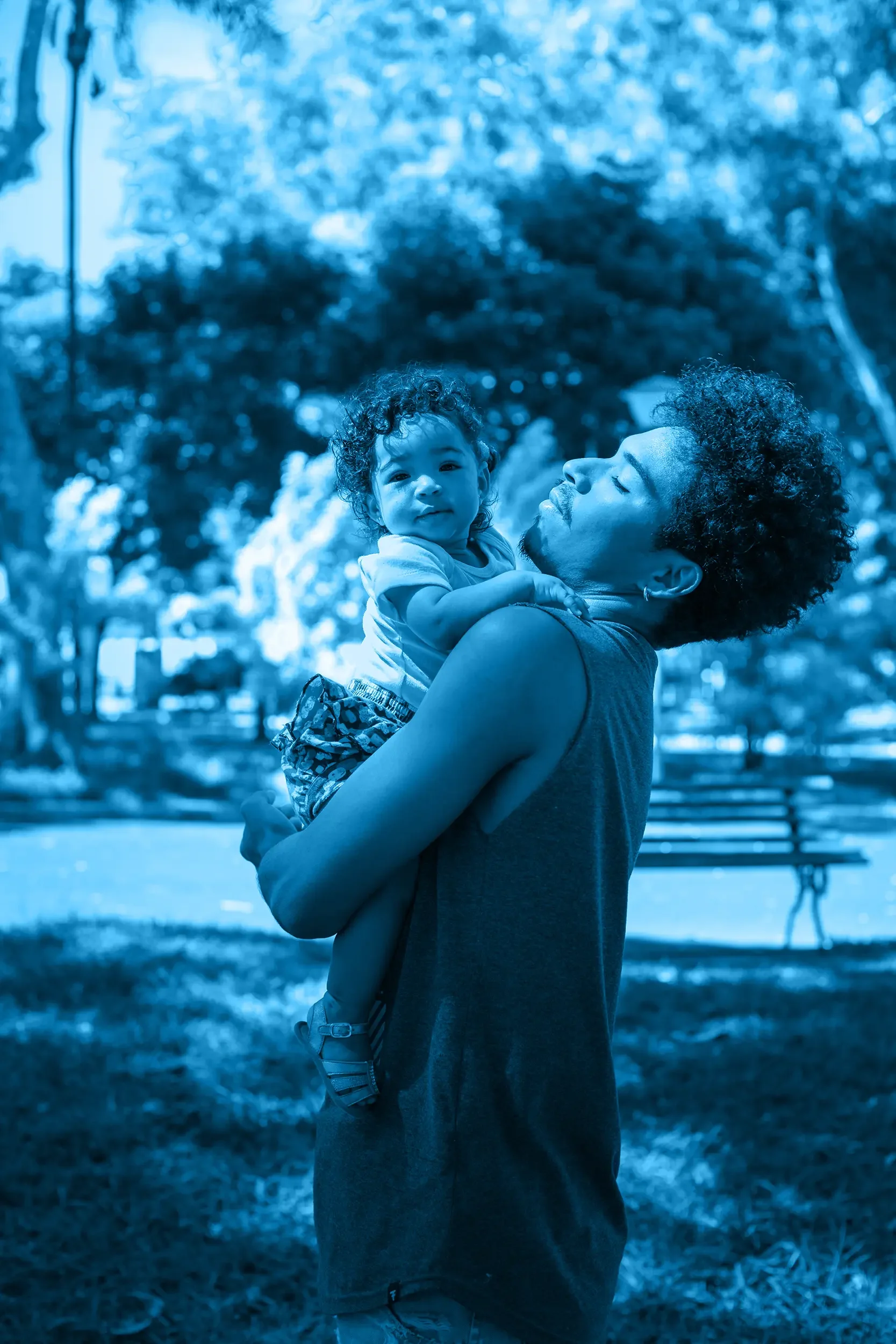 A dad with curly hair holding a young child with curly hair in a park with trees and a bench in the background.