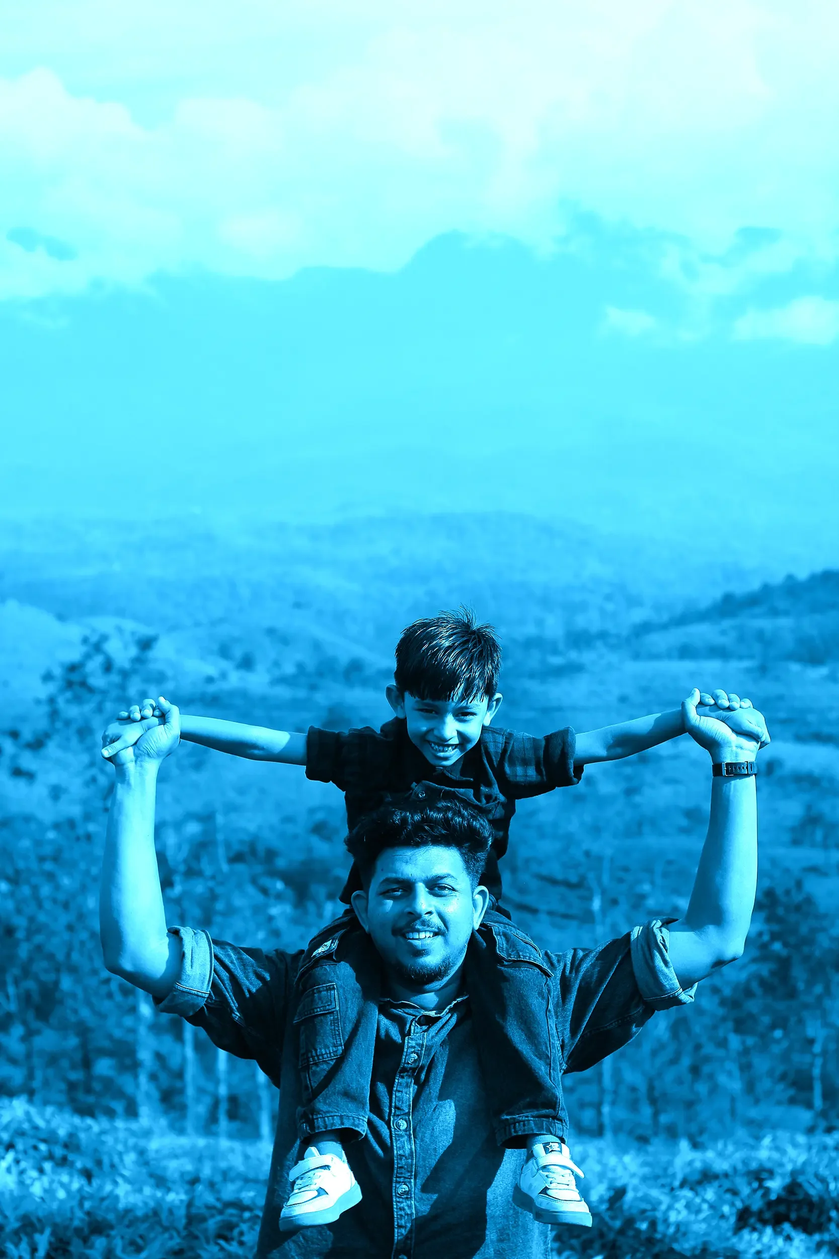 A father with a young boy on his shoulders on a grassy hillside with a scenic view of mountains and clouds in the background