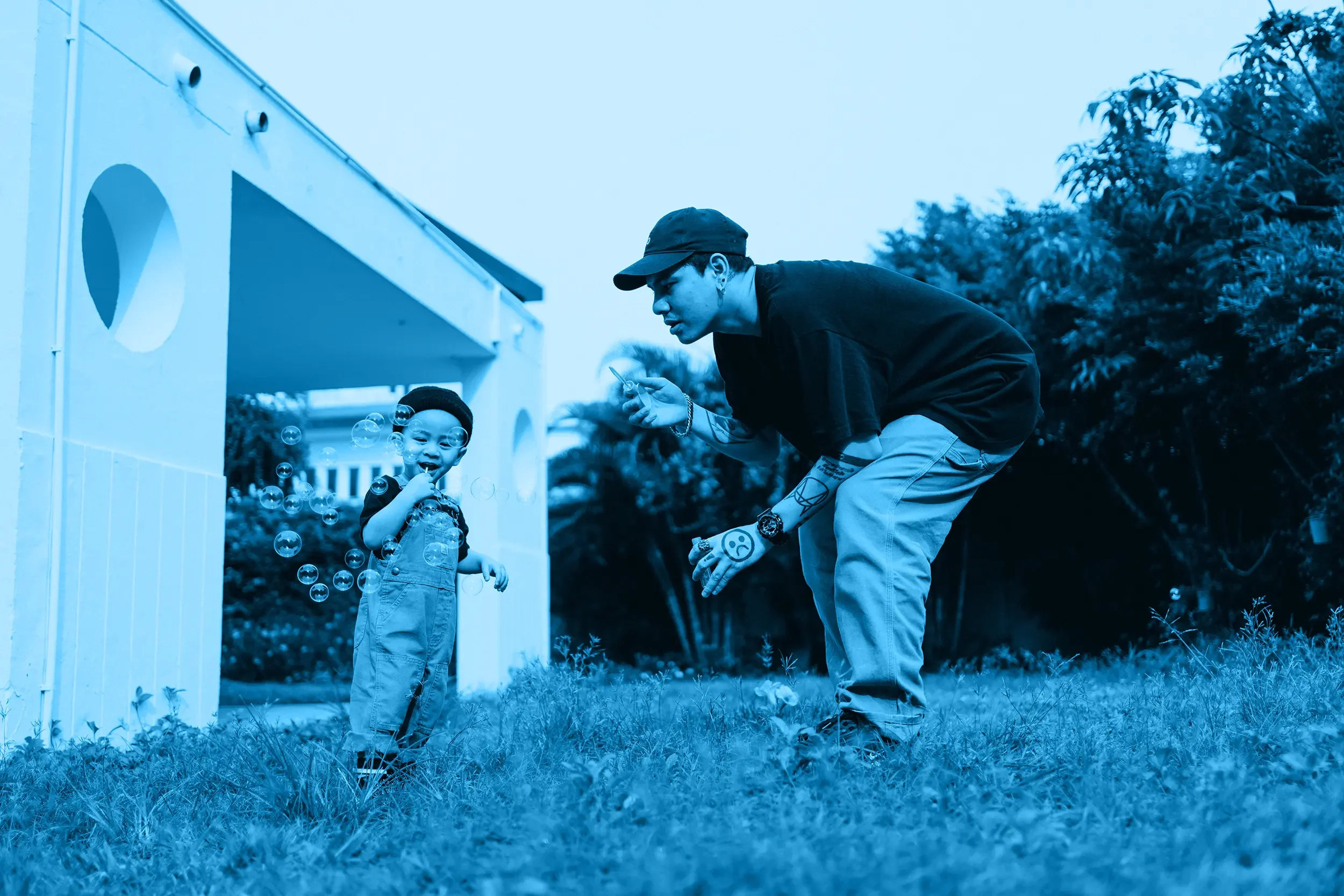 A man with tattoos and a cap is playing with a young boy in a backyard, blowing bubbles.