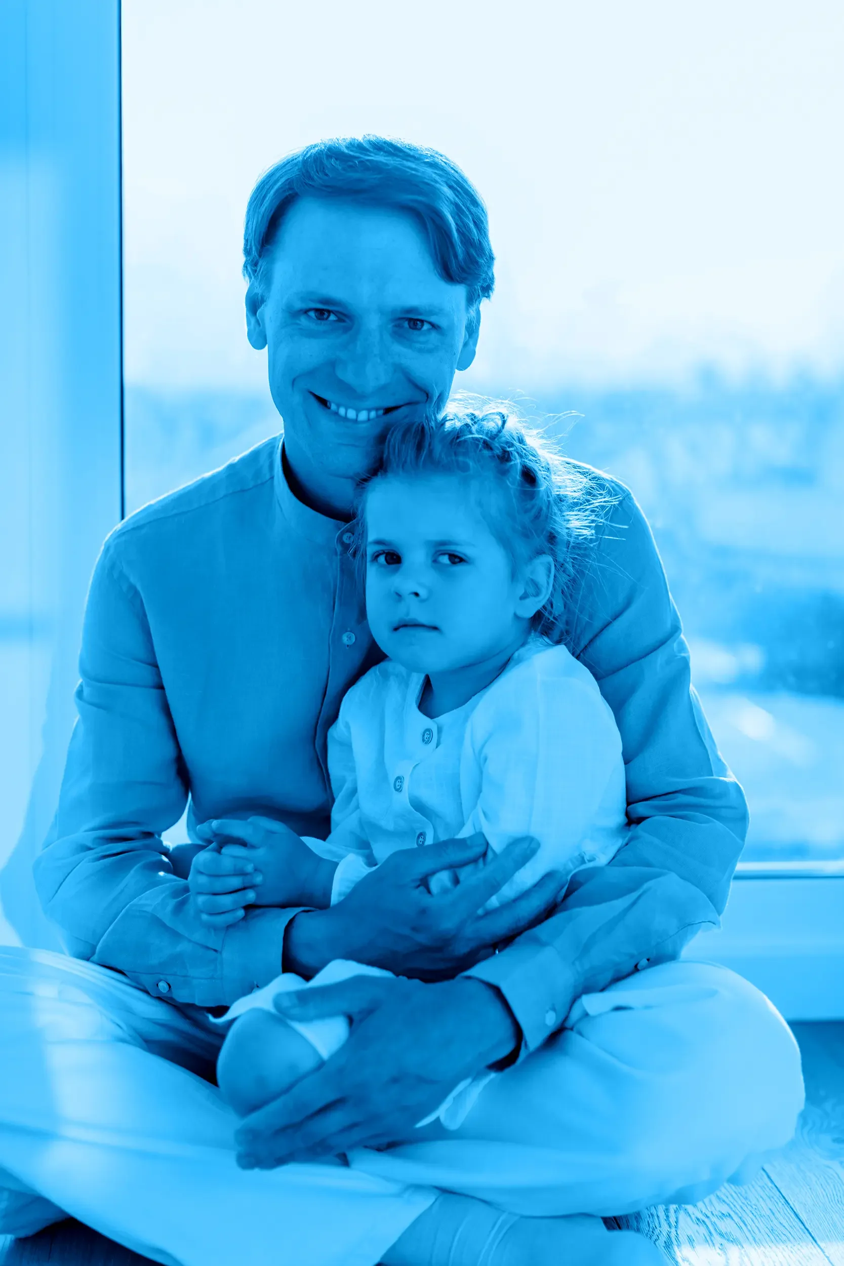 A father and a young girl sitting together near a window, with a blue color overlay. The man is smiling and the girl looks serious.