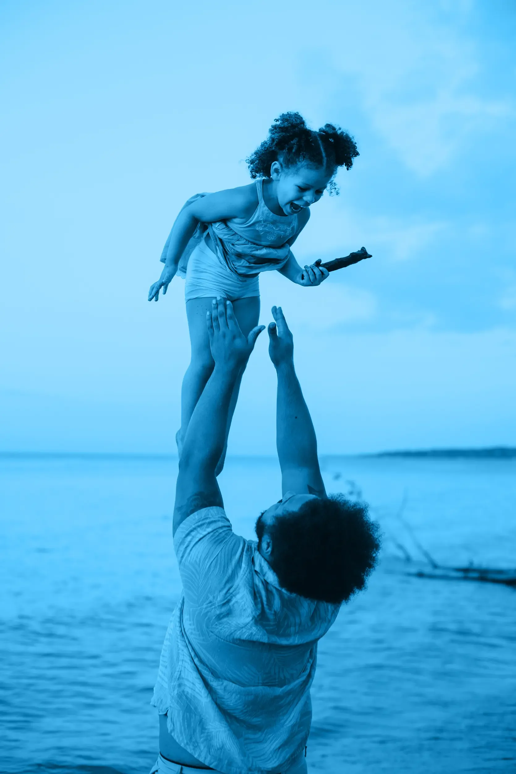 A father at the beach lifting a young girl in the air. The girl is holding a stick and appears to be shouting or laughing. The background shows the ocean and sky.