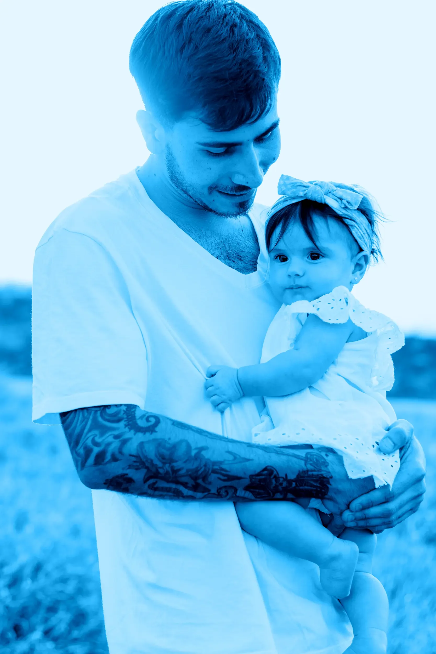 A dad with tattoos on his arm holding a young girl, both smiling and looking down. The girl wears a headband with a bow. They are outdoors with a blurred natural background.