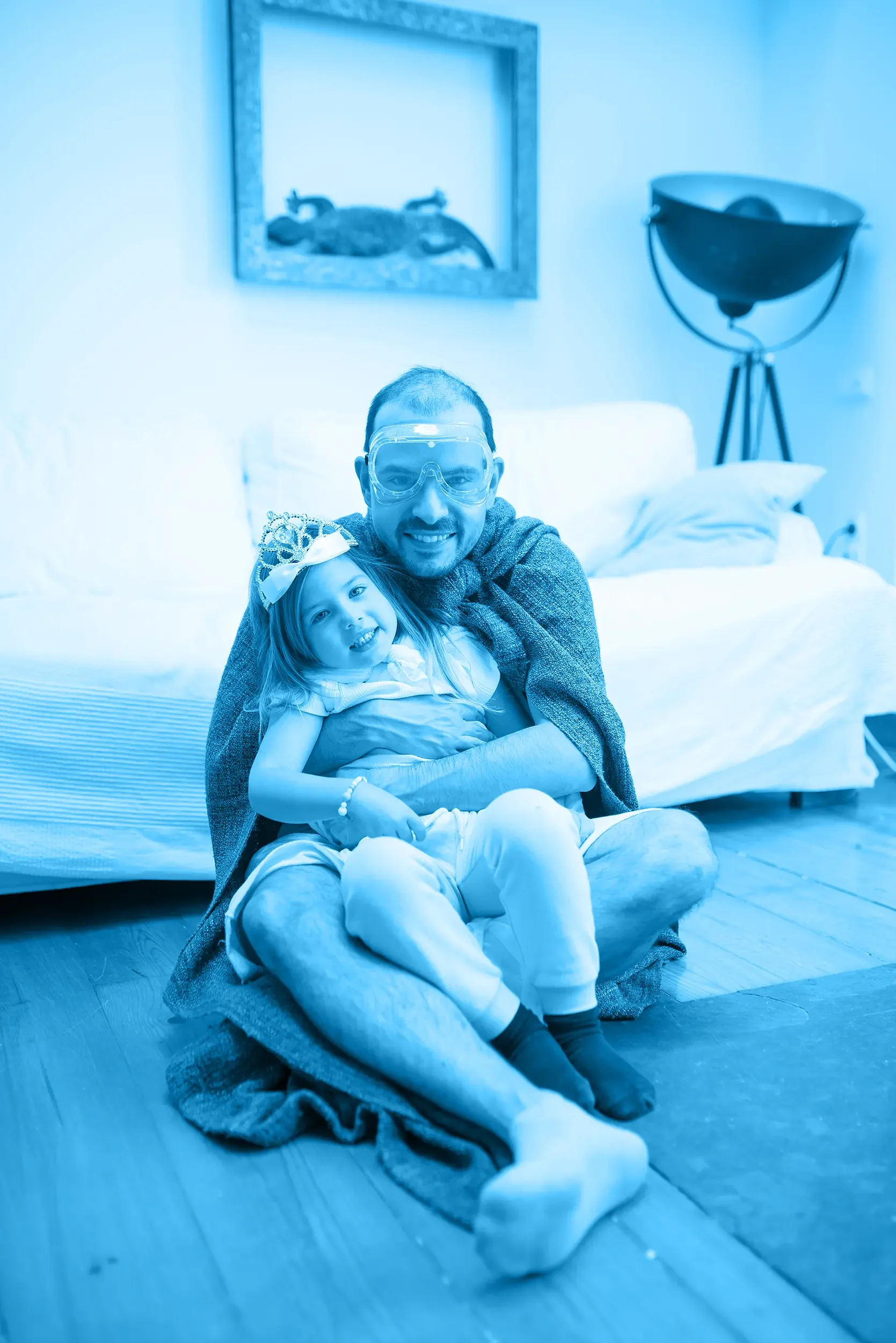 A father and a young girl sitting on the floor of a living room, smiling at the camera. The girl is wearing a large bow on her head, and the man is wearing glasses. Behind them is a bed and a framed picture of a lizard on the wall.